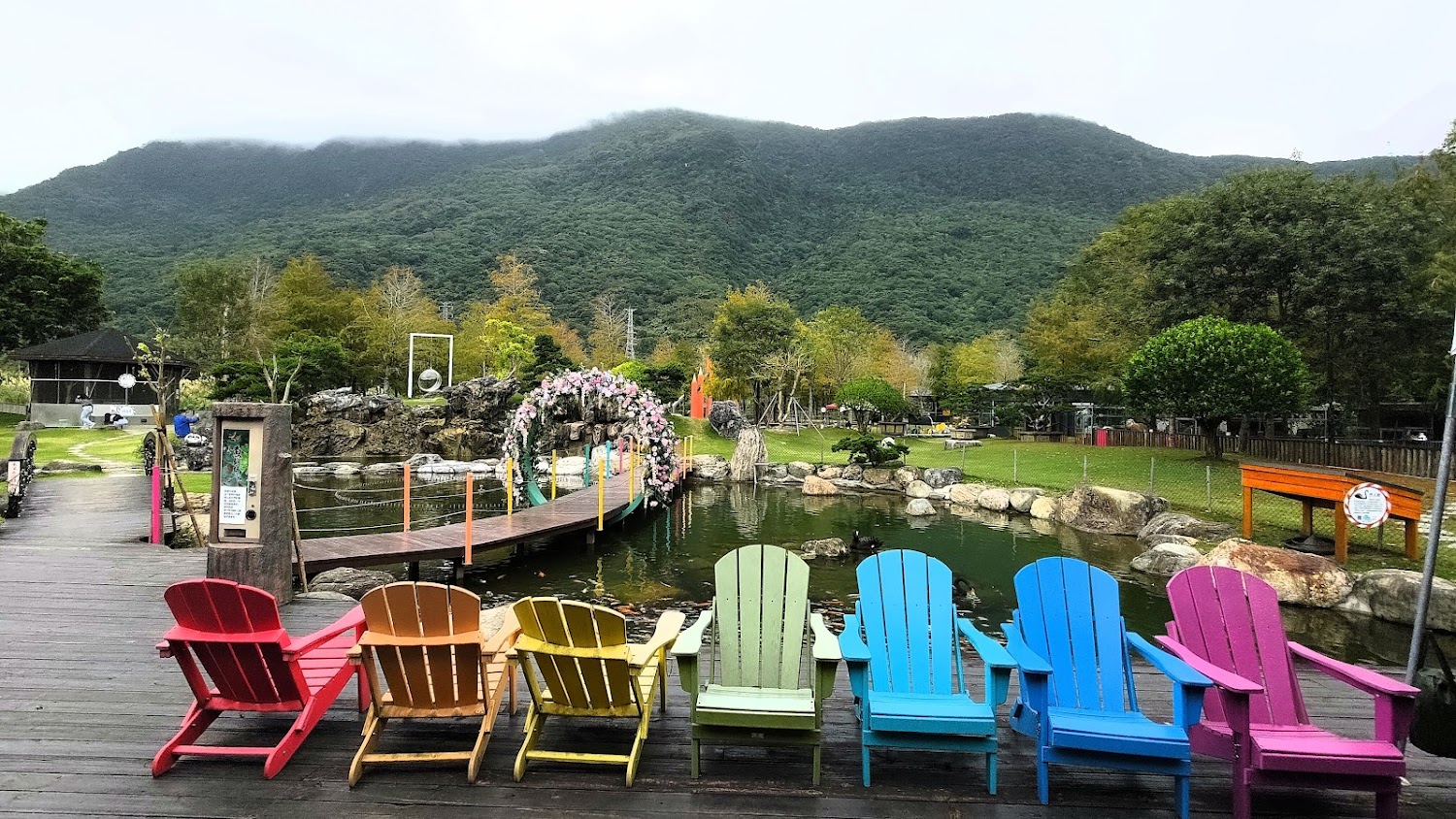 七張彩色椅子面向池塘與花圈木橋，背景是綠色山林