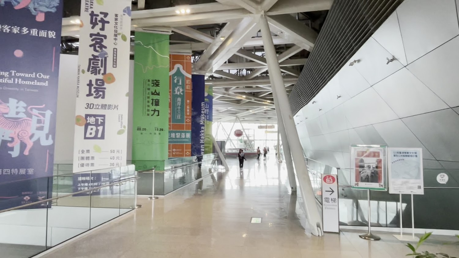 這是一個現代化博物館或展覽館的室內空間，左側有大型展覽布條與標示，地板明亮乾淨，玻璃扶手與開放式空間設計，右側有通往電梯的指示牌，通道盡頭有人走動。
