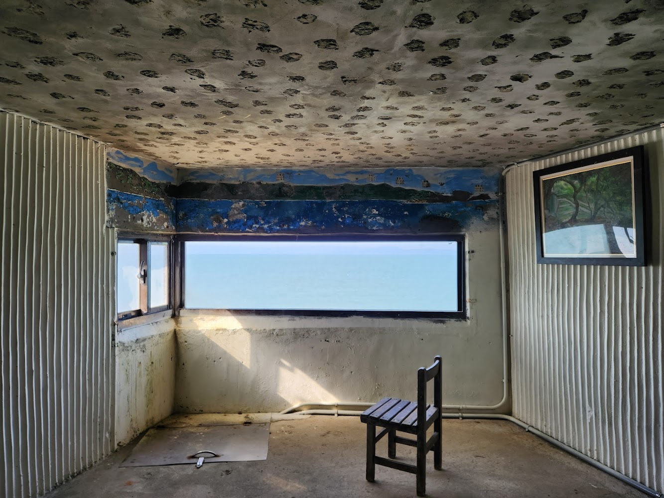 這張圖片主要展示一間老舊房間，牆上有窗戶可以看到海景，室內有一張簡單木椅和一幅掛畫，天花板有特殊的裝飾圖案。