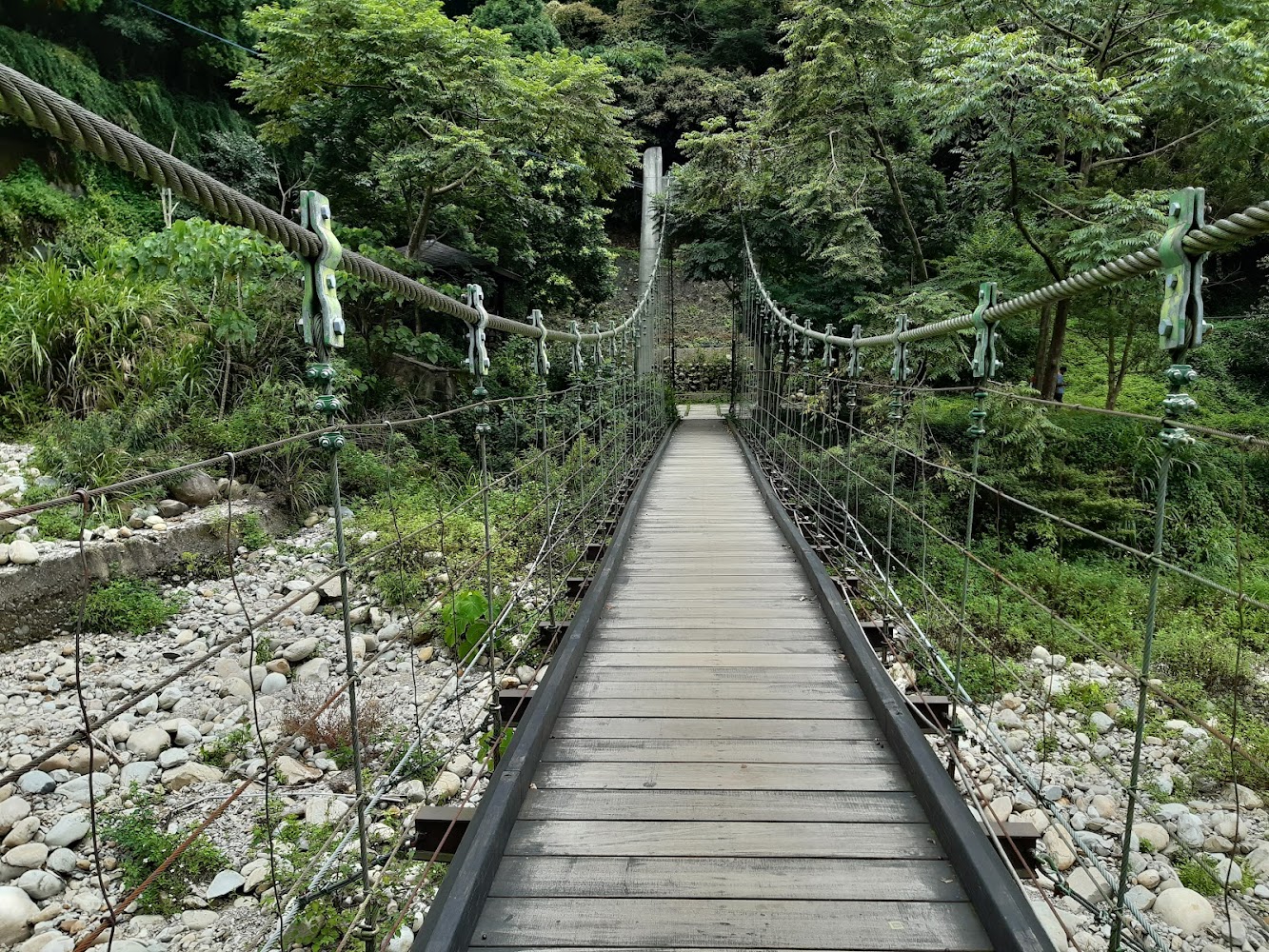 這張圖片主要是一座橫跨河谷的吊橋，橋面由木板構成，兩側用鋼索與鐵絲網支撐，四周環繞著茂密的綠樹與石頭河床，展現大自然的美景。
