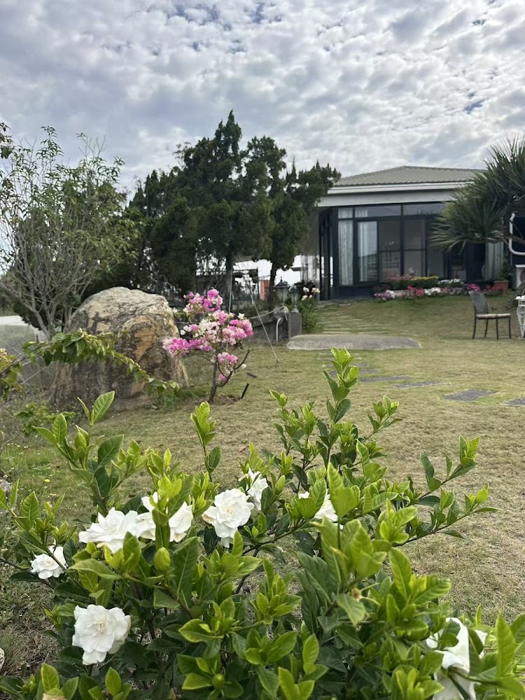 這張圖片展示一個庭院，前景有盛開的白色花卉與綠色植物，中間有大石頭和粉紅色花，遠處有一間現代感的玻璃屋和樹木，環境自然寧靜。圖中無明顯浮水印。
