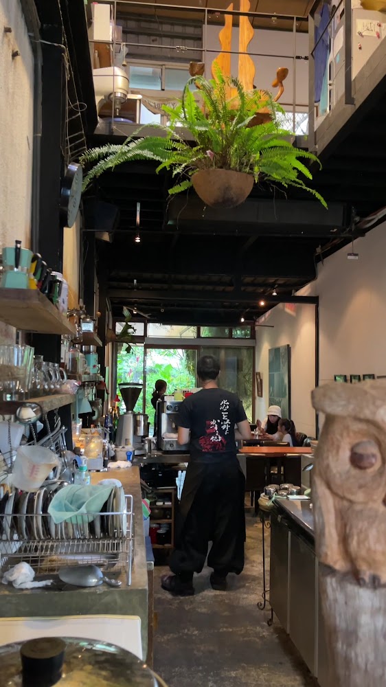 這是一間有著植物裝飾的小型咖啡館，吧台上有各式器具與餐具，裡面有工作人員及幾位客人在用餐，環境溫馨且帶有自然氛圍。