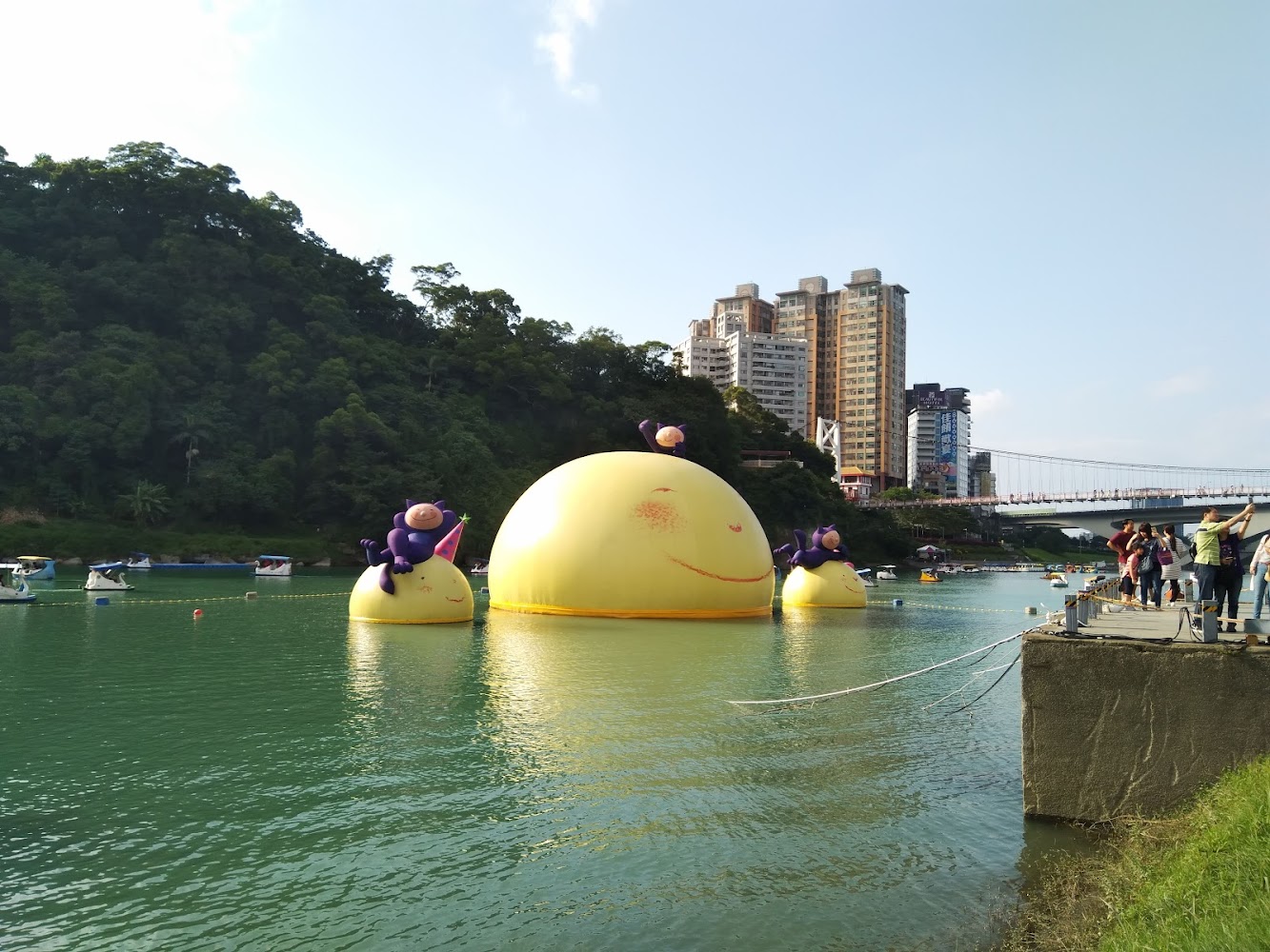 湖面上漂浮大型黃色充氣裝置與紫色卡通角色，背景有高樓與吊橋
