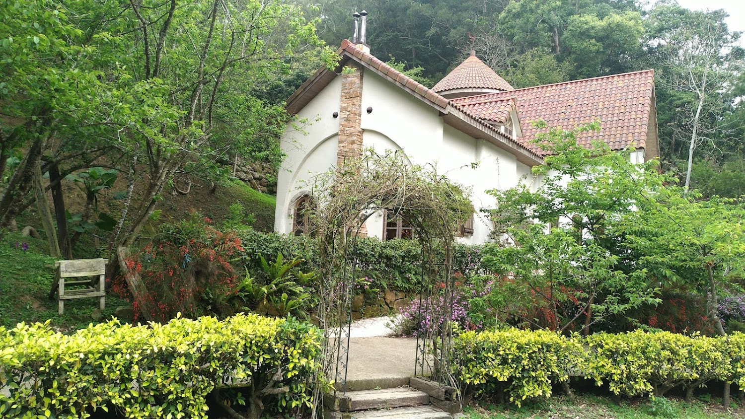 這張圖片主要是一棟帶有圓錐屋頂的歐式小屋，四周被茂密的樹木、花草和綠色灌木環繞，前方有一座藤蔓拱門與小徑，環境十分幽靜優美。