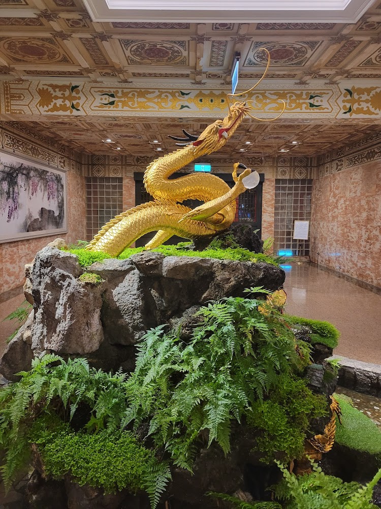 一條金色中國龍雕塑,盤踞在佈滿綠色蕨類植物的假山上,背景為華麗的裝飾天花板及牆面