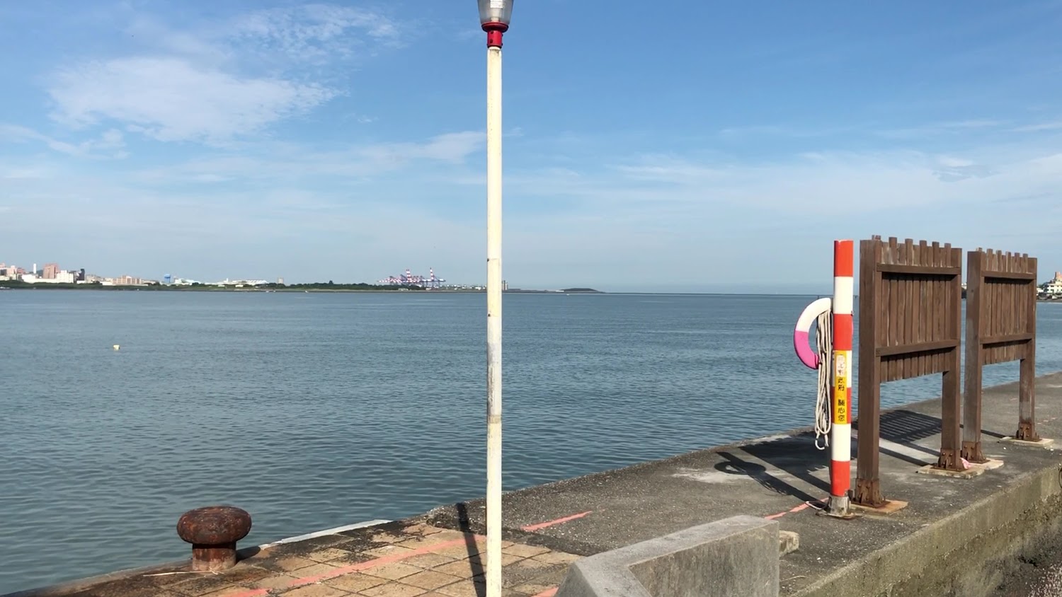 海邊碼頭有燈桿、救生圈、木質護欄與清澈海面，遠方可見城市建築