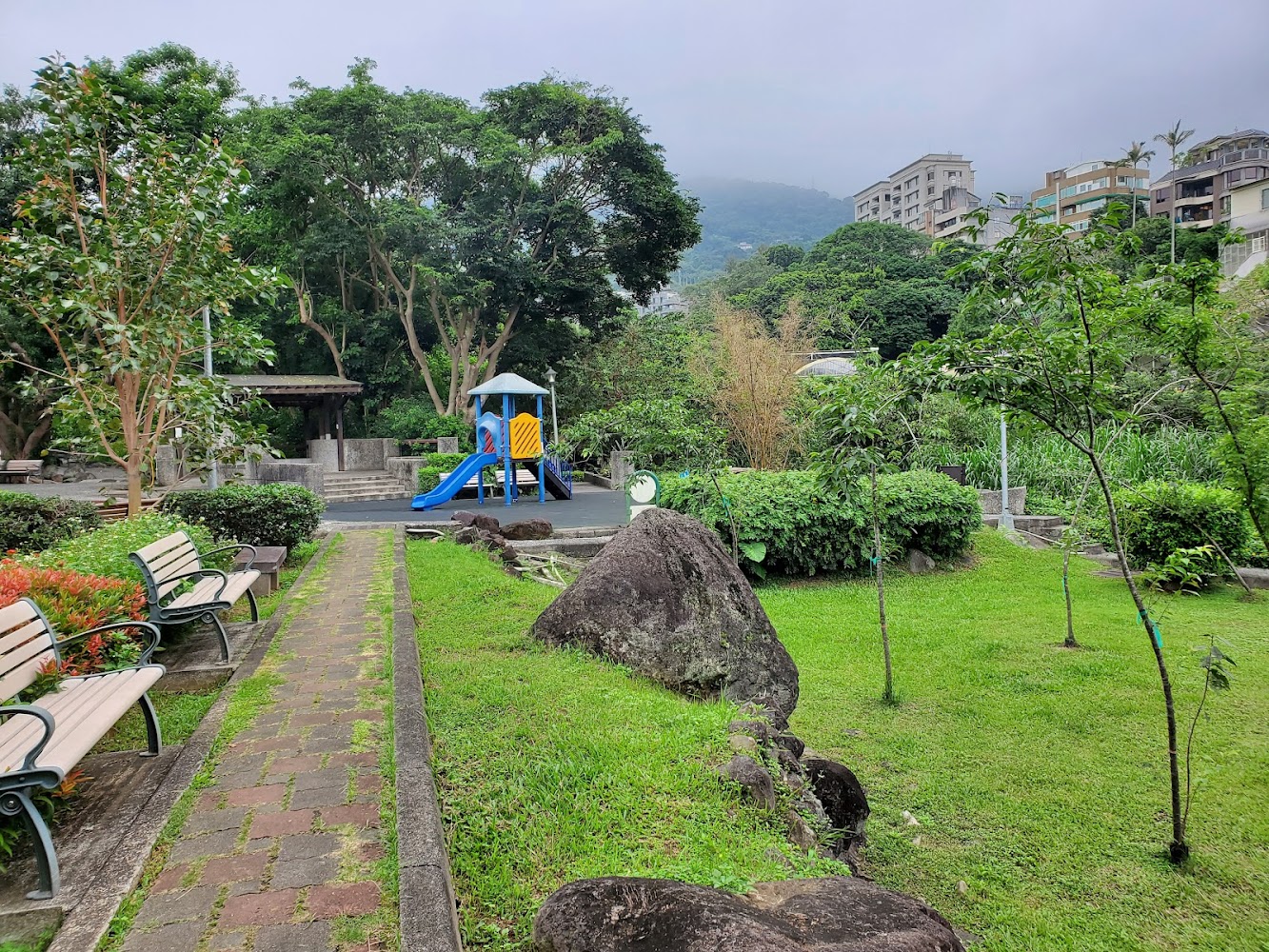 社區公園有長椅、兒童遊樂設施、草地、樹木與岩石，背景有山林及住宅