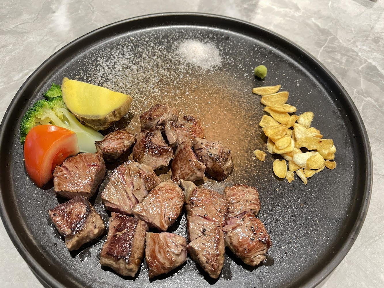 這張圖片主要是鐵板牛排丁，搭配烤蒜片、鹽調味、芥末、馬鈴薯、花椰菜和番茄，擺盤精緻美觀。