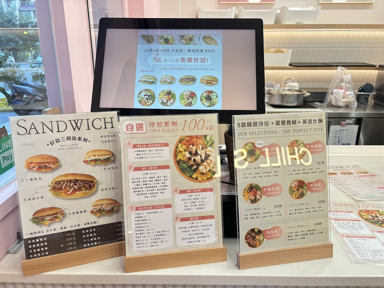這張圖片主要展示一家輕食店的點餐櫃檯與菜單，包含三明治、沙拉與各式套餐選擇，並有活動宣傳資訊，風格明亮整潔。
