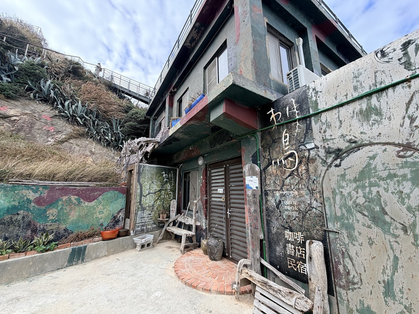 這張圖片主要展示一間外觀充滿藝術塗鴉風格的建築，牆上有咖啡、書店、民宿等字樣，門口擺放木製座椅，旁邊則有植物與山坡，環境自然又具特色。