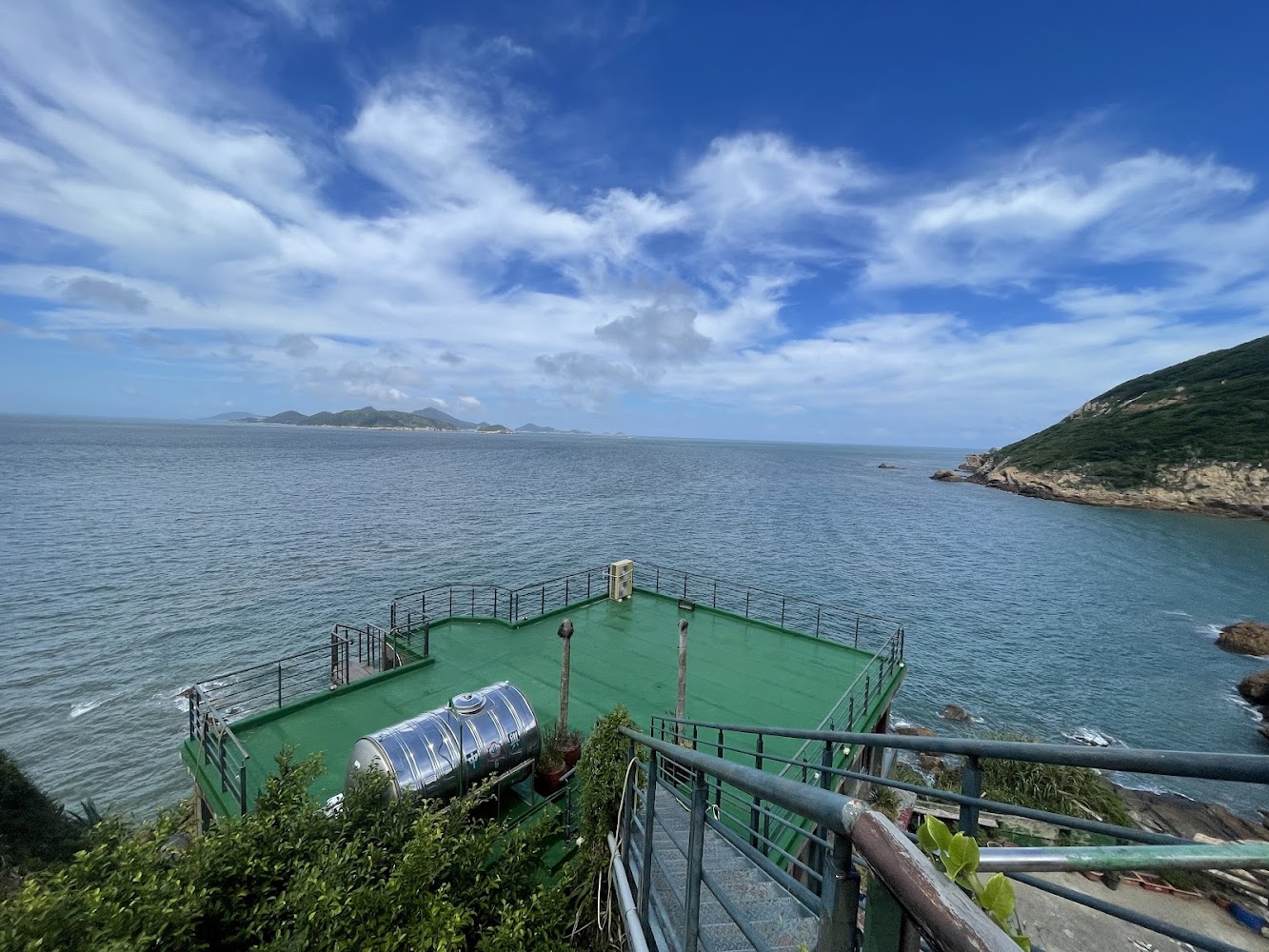 這張圖片主要展示一片海景、藍天白雲，右側有山丘。前方有一個綠色平台，平台上有一個不鏽鋼水箱，左下角有樓梯和植物。