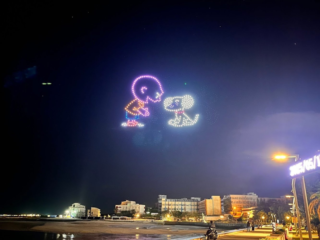 夜空中的無人機燈光秀組成小孩與小狗圖案，下方是城市夜景。