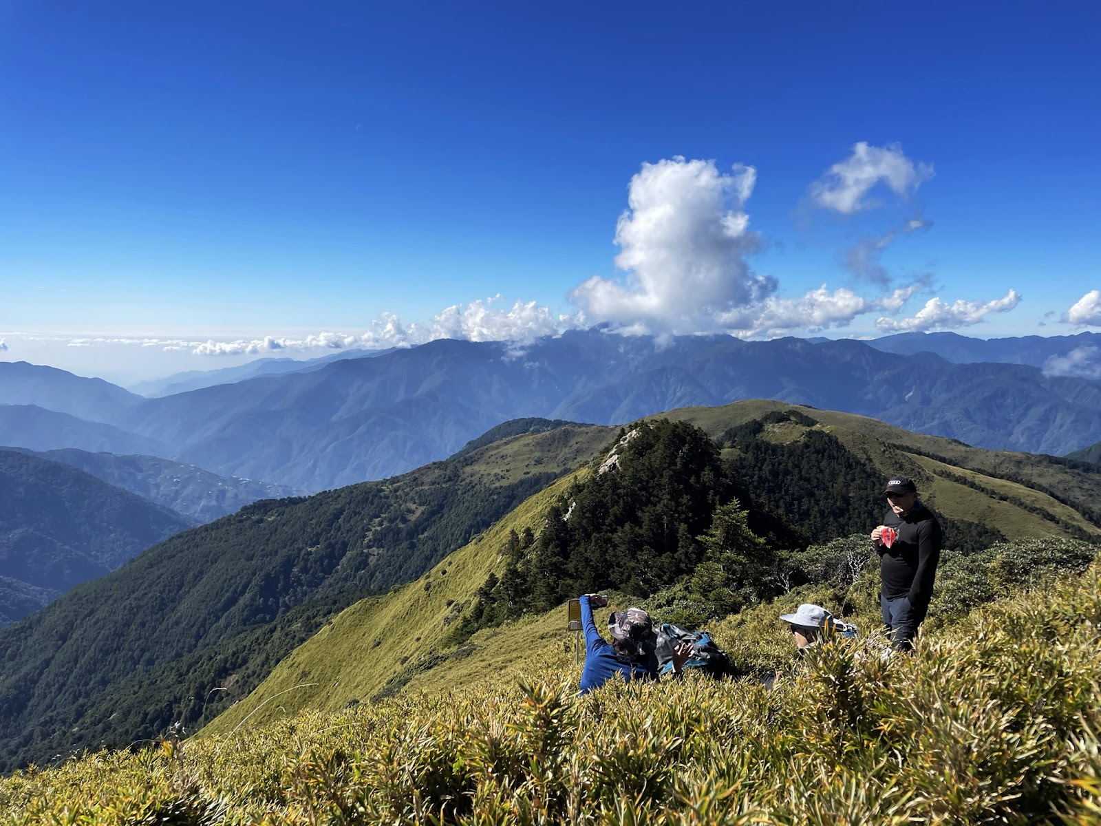登山客在合歡山草坡山頂休憩，一人臥地拍照，背景翠綠稜脊與藍天白雲