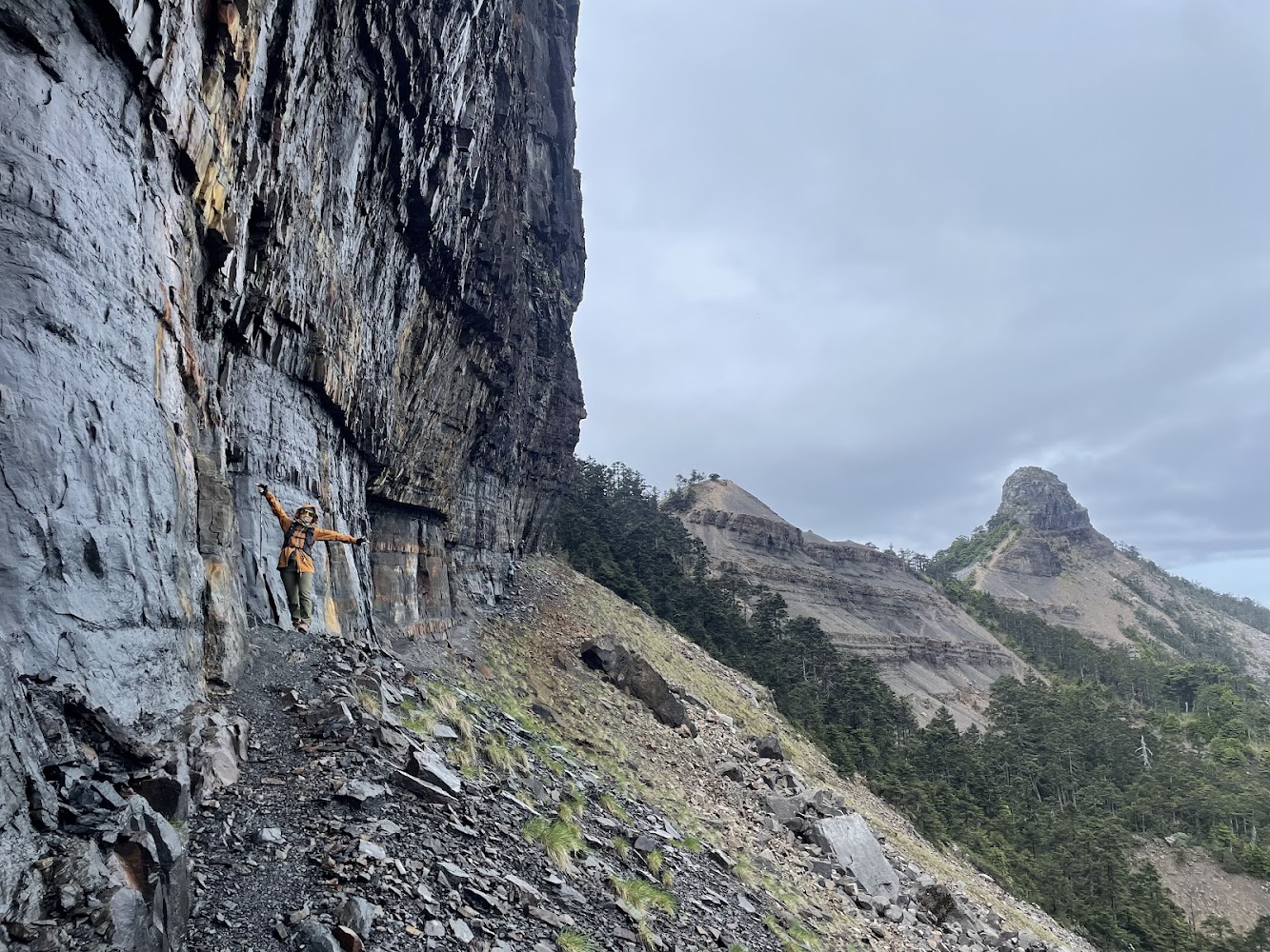 穿橘色外套的登山者站在岩壁邊遠望山林