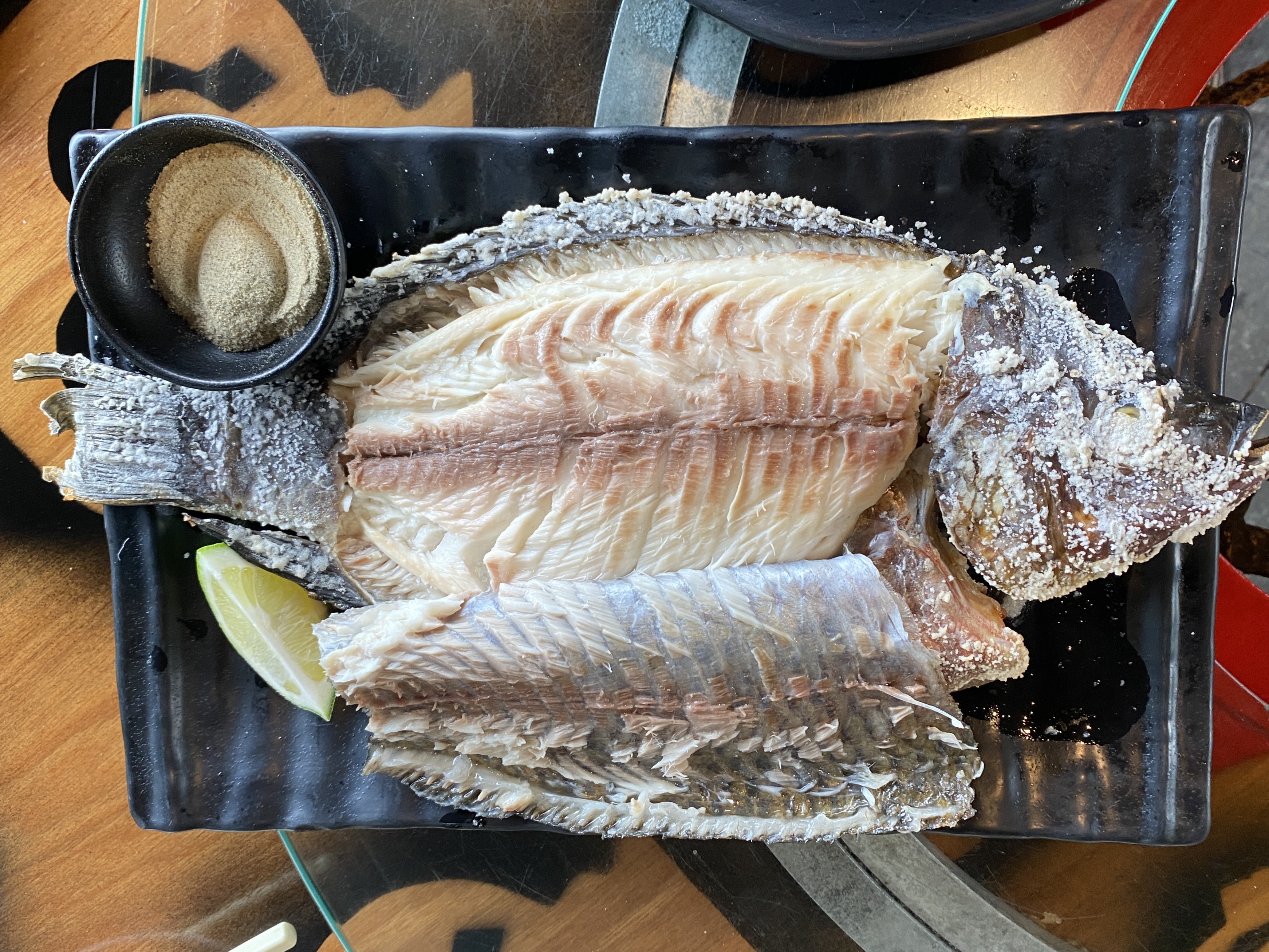 一道鹽烤魚，魚身劃開，搭配檸檬以及胡椒鹽，擺放在黑色長盤上