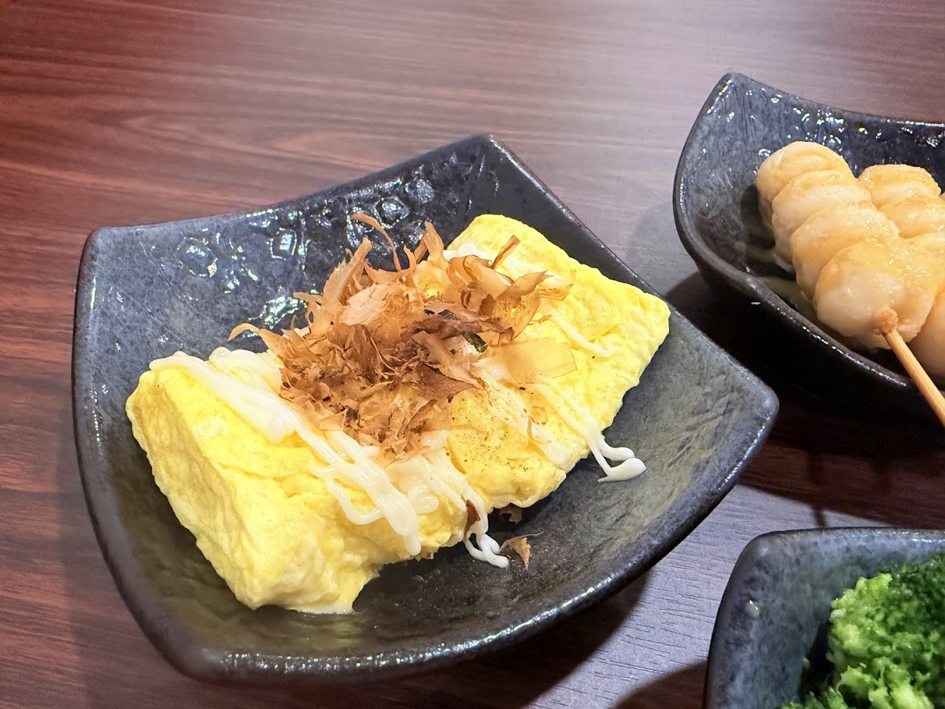 這張圖片主要有一道日式玉子燒，上面放有柴魚片和美乃滋，旁邊還有串烤糯米糰和一點青菜，擺放於黑色餐盤上。