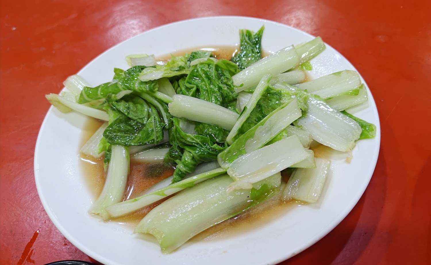 這張圖片是一盤炒青菜，青菜被盛裝在白色圓盤中，底下有醬汁，看起來清爽健康，適合搭配主餐享用。