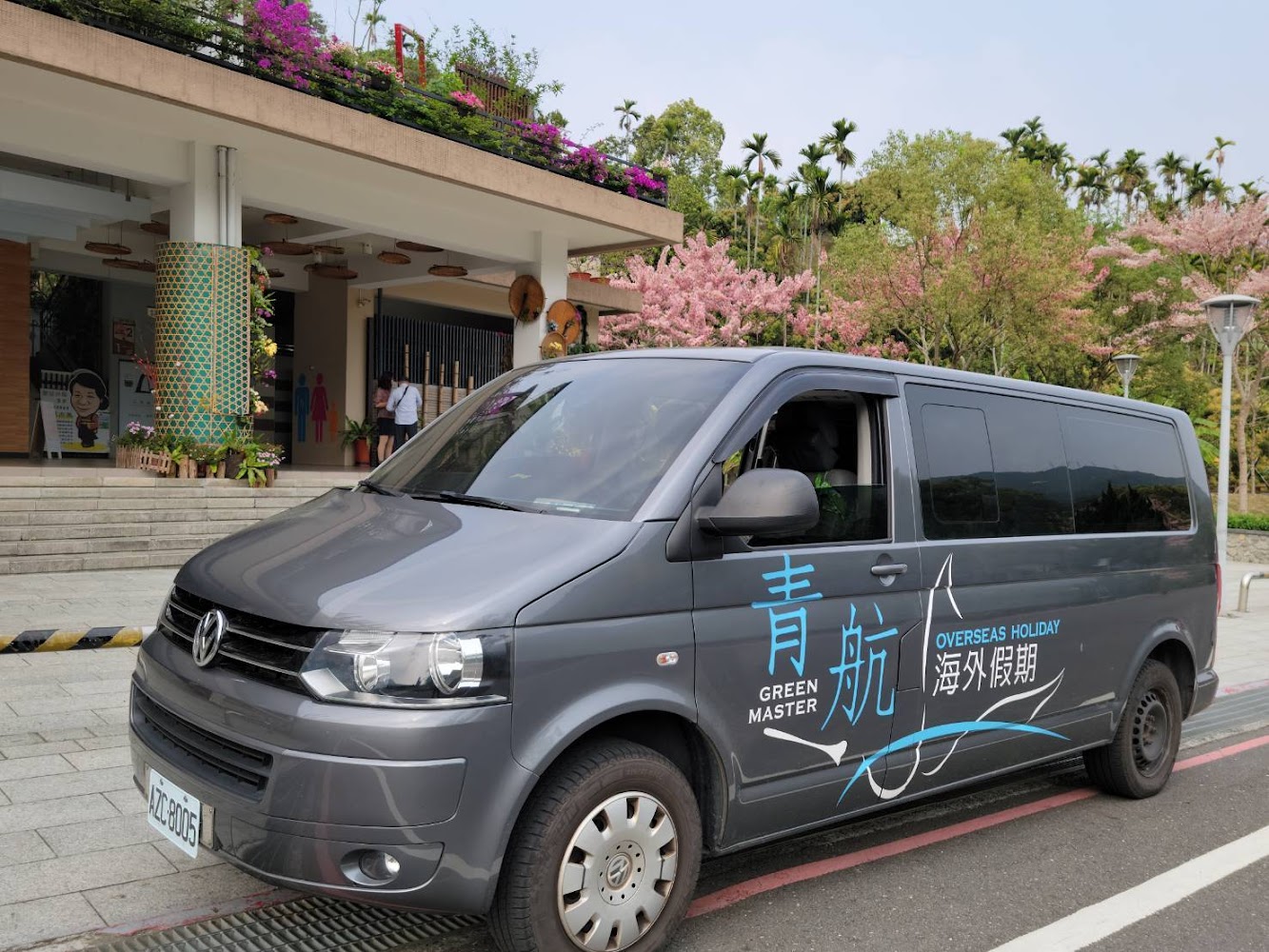 停在建築物前的深灰色廂型車，背景有花卉和綠樹