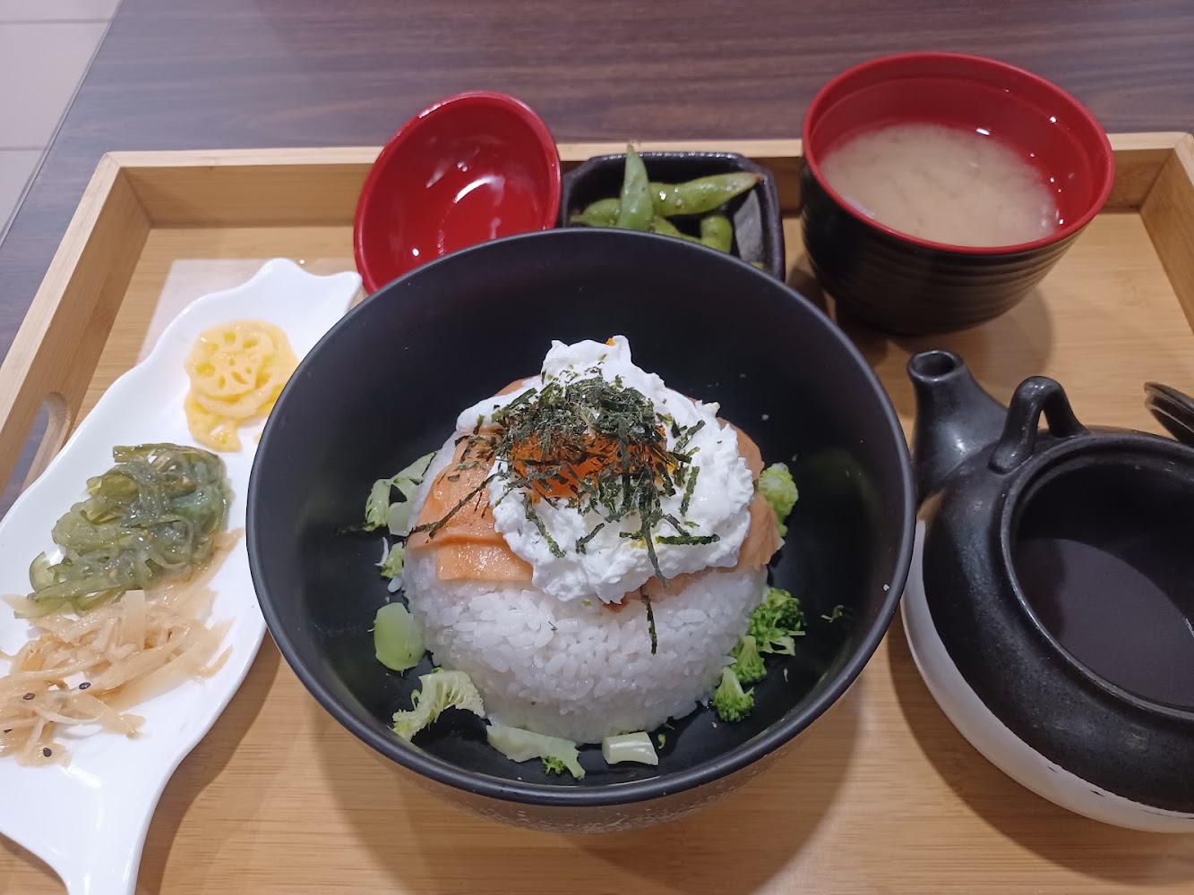 這是一份日式套餐，包括一碗鮭魚飯上覆有魚卵、海苔和醬料，旁邊搭配味噌湯、小菜與毛豆，以及一壺茶湯可沖飯食用，擺盤精緻豐富。