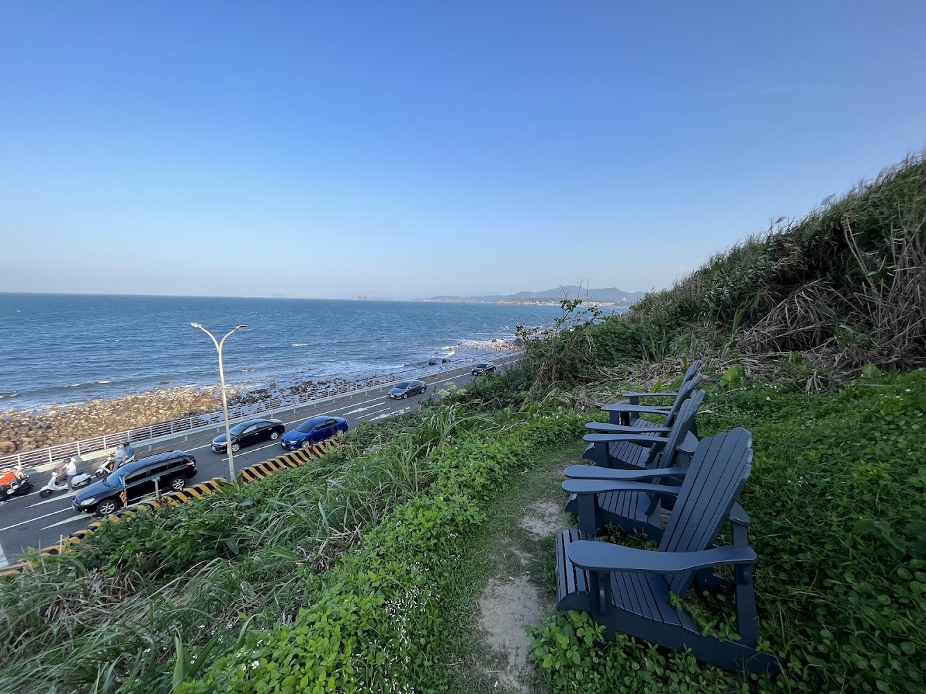 草地上排列的休閒椅前方是沿海公路與藍色海洋