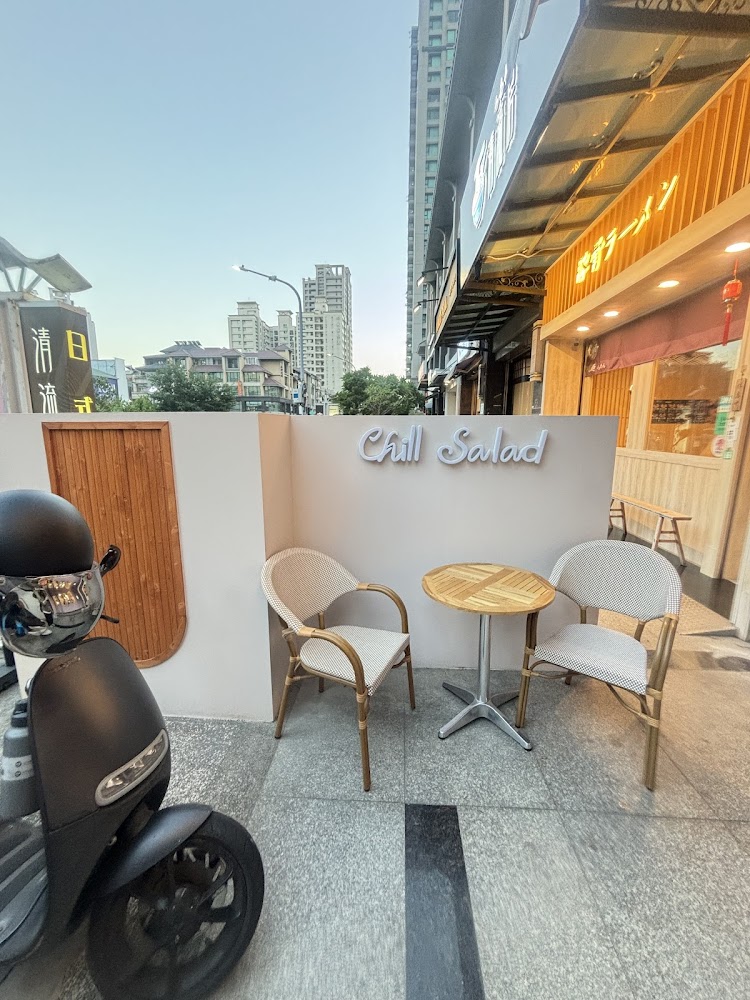 這張圖片展示了一家名為Chill Salad的店門口，門前有兩張椅子、一張圓形木桌，旁邊停著一台黑色機車。背景還可見高樓與街景。