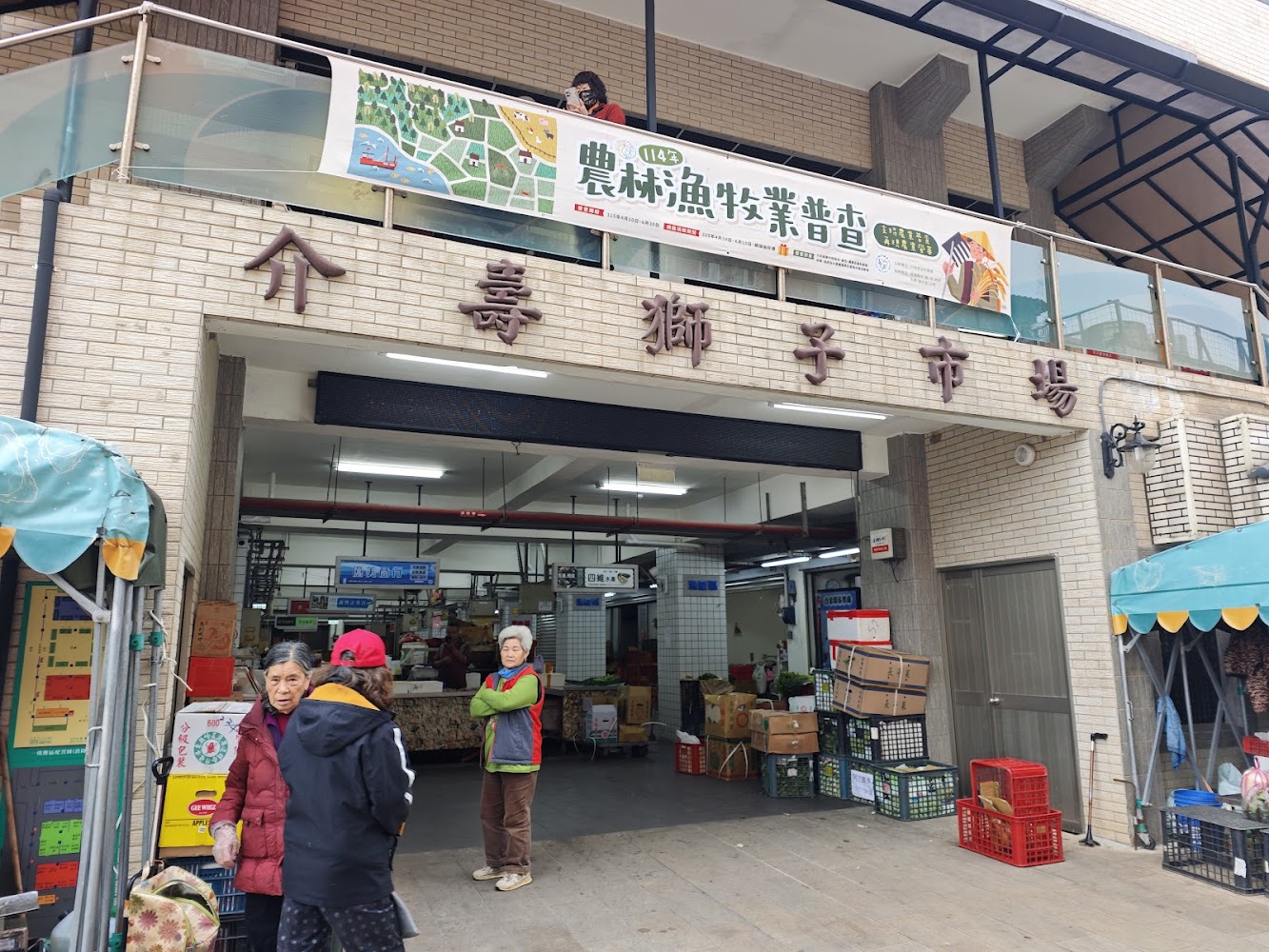 這是一個傳統市場的入口，招牌寫著『壽獅子市場』，門口有幾位長者和居民，周邊有攤販、貨物箱與蔬果，展現熱鬧的在地生活氛圍。