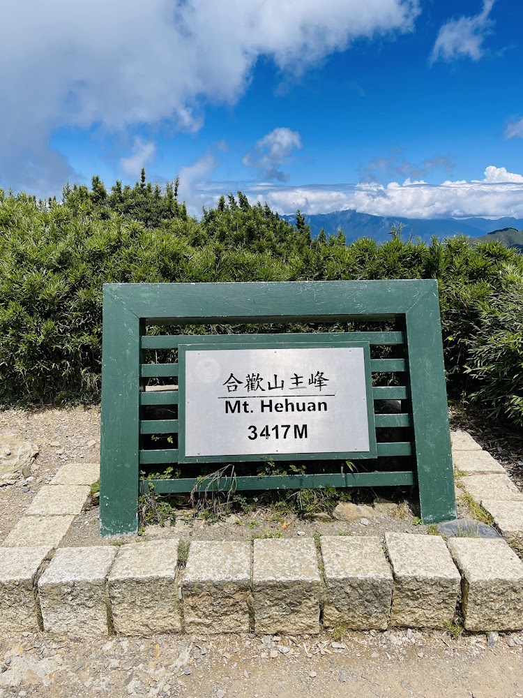圖片主要是一座標示牌,寫著『合歡山主峰 Mt. Hehuan 3417M』,背景有藍天、白雲和綠色山景。圖片沒有浮水印。