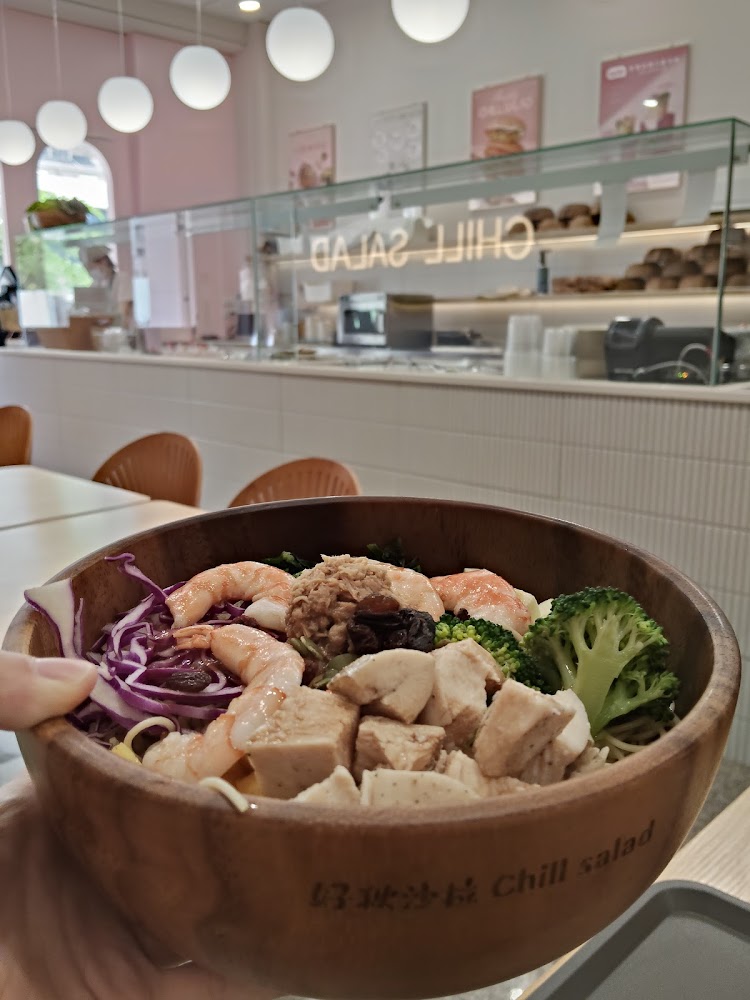 這張圖片展示了一碗豐富的沙拉，內含雞胸肉、蝦仁、鮪魚、葡萄乾、紫甘藍和花椰菜，背景為裝潢現代的沙拉店。