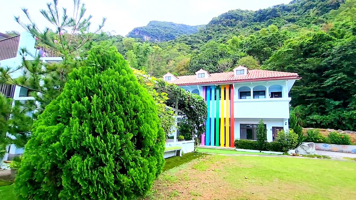 白色兩層樓建築外牆有彩色直條紋，前方是整齊草坪與樹木，背景為山林