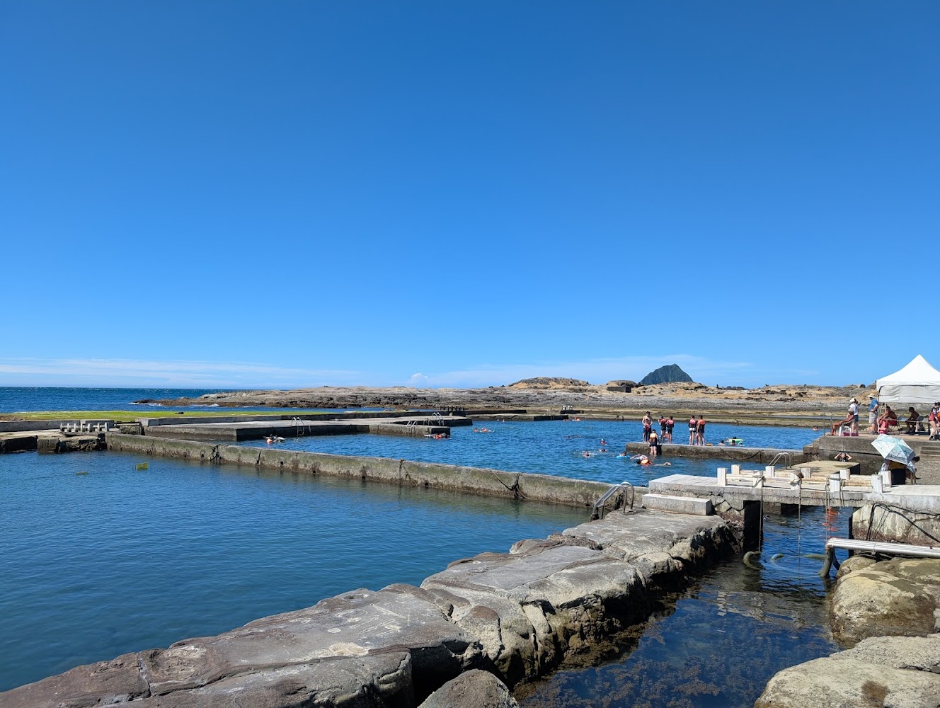 人工建造的海水泳池，池中有人戲水，背景是大海與藍天
