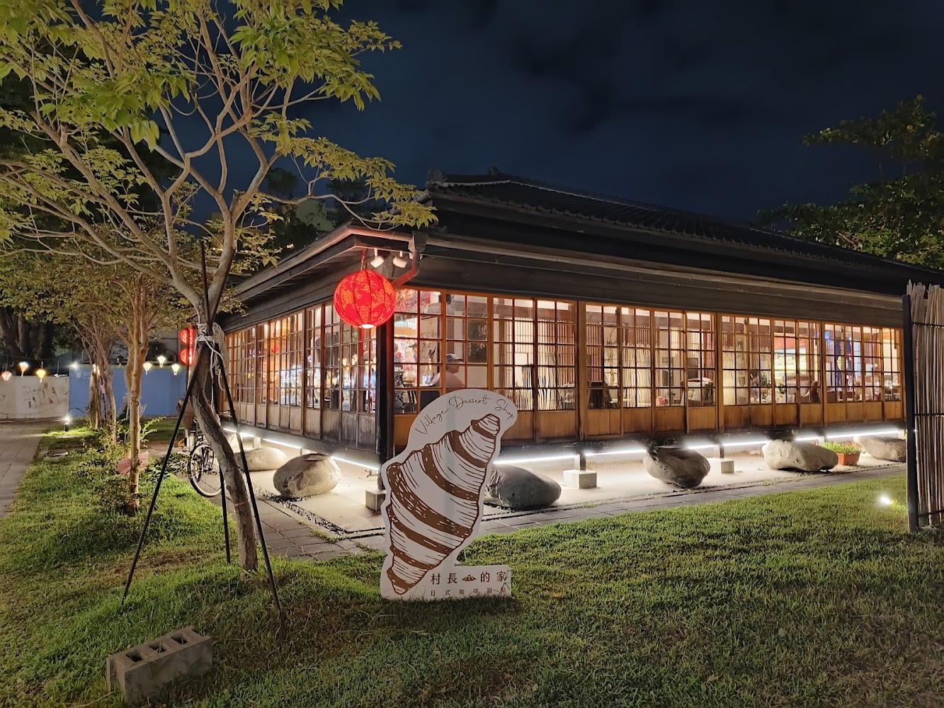 夜晚時分的木構日式建築，玻璃窗透出室內燈光，外有紅燈籠與草地