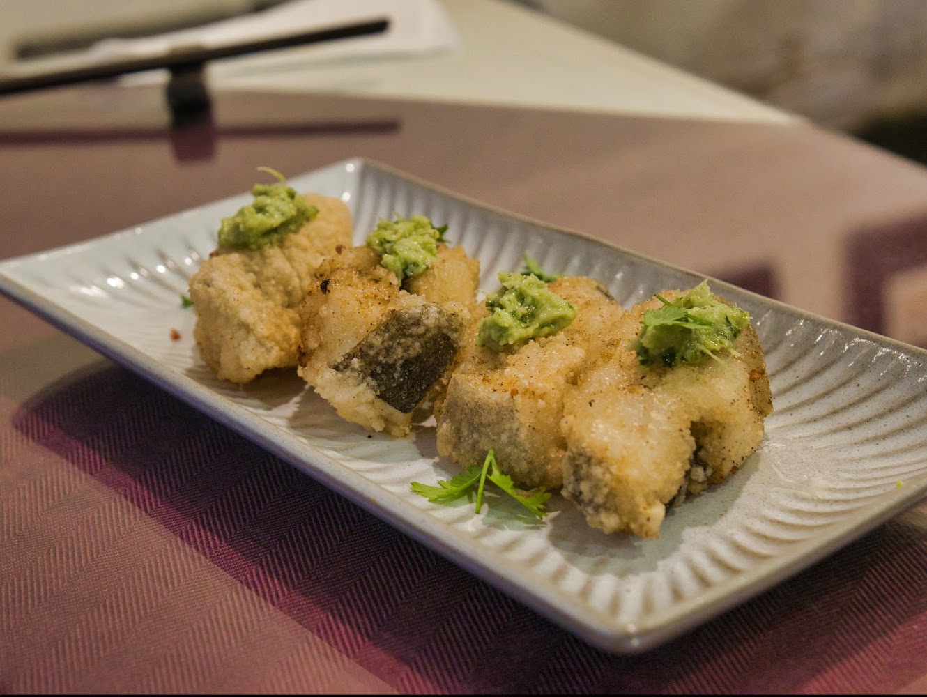 這張圖片主要呈現一道日式料理，盤中有四塊裹粉炸魚塊，魚塊上方各放有綠色醬料和香菜葉，擺放整齊於陶瓷長盤中。