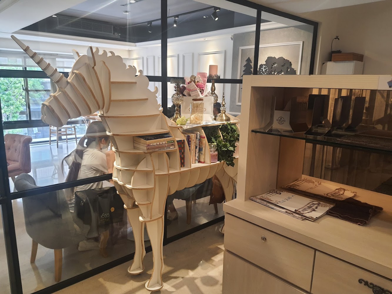 這張圖片主要展示一個木製的獨角獸書架，上面擺放著書籍與裝飾品，背景則是一家明亮、現代風格的咖啡廳，內有人正在用餐與交談。
