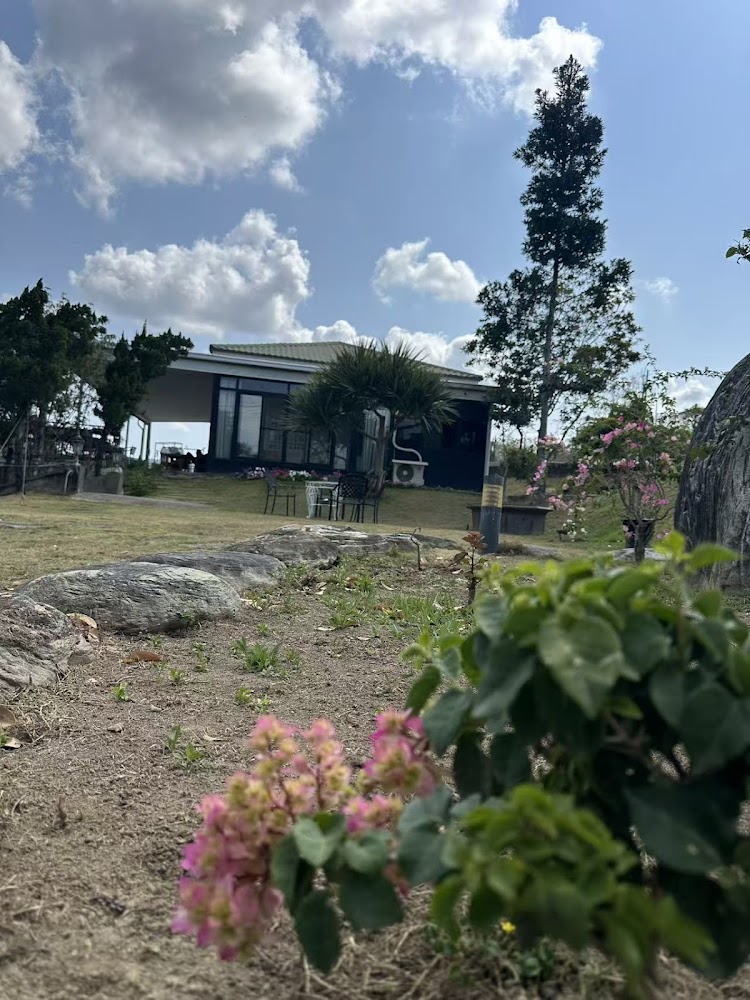 這張圖片展示了一棟現代感的小屋，前方有綠地及庭院，搭配幾張戶外桌椅。地面有石頭及花叢，天空晴朗、白雲飄浮，景色寧靜自然。圖片上未見浮水印。