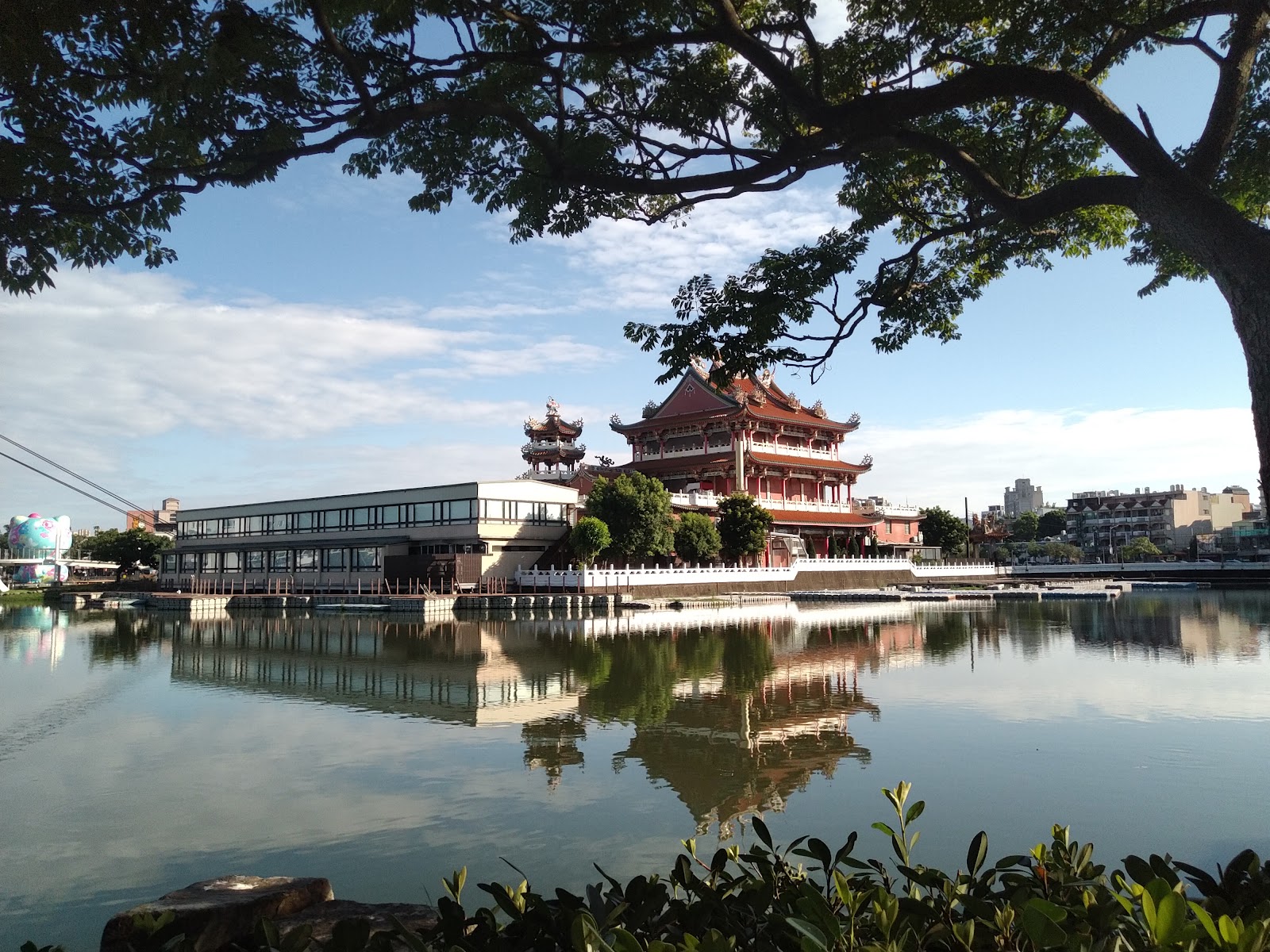 龍潭大池白天，紅色中式廟宇倒映水中，大樹枝葉框景，藍天白雲