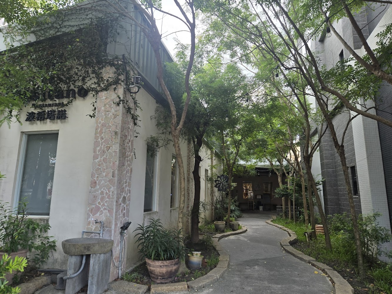 圖片中有一家名為『波希若諾』的餐廳，建築外牆有藤蔓和石材裝飾。旁邊有洗手台與多個花盆，沿著小徑兩旁種植著綠樹，環境自然清幽。