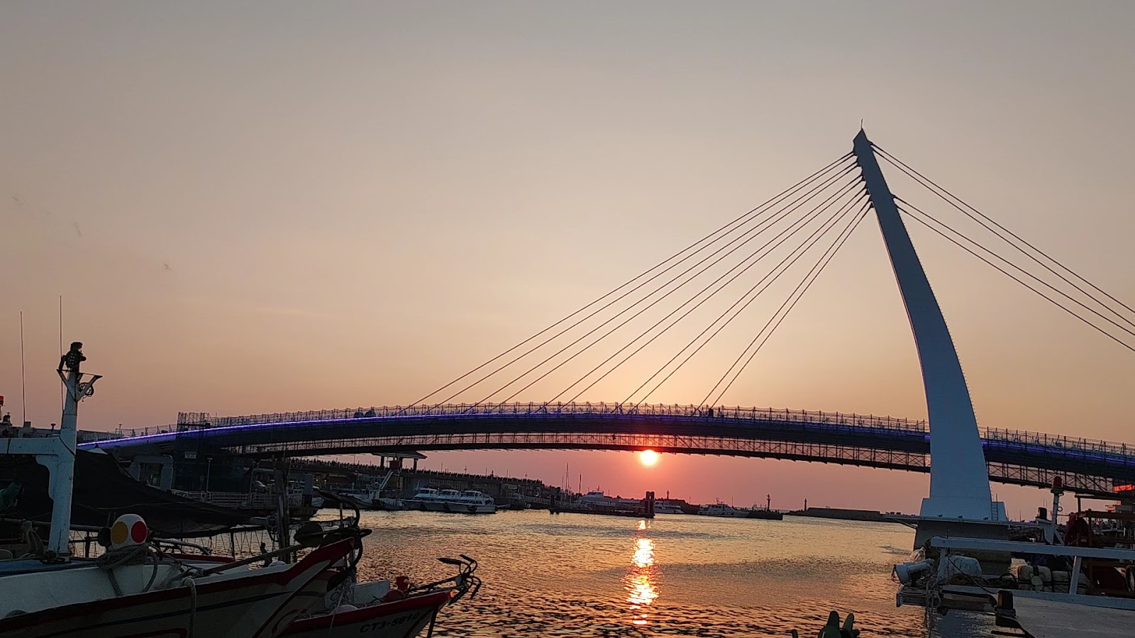 情人橋夕陽剪影，橋身藍紫燈光，夕陽映水金橙色倒影，前景漁船停泊