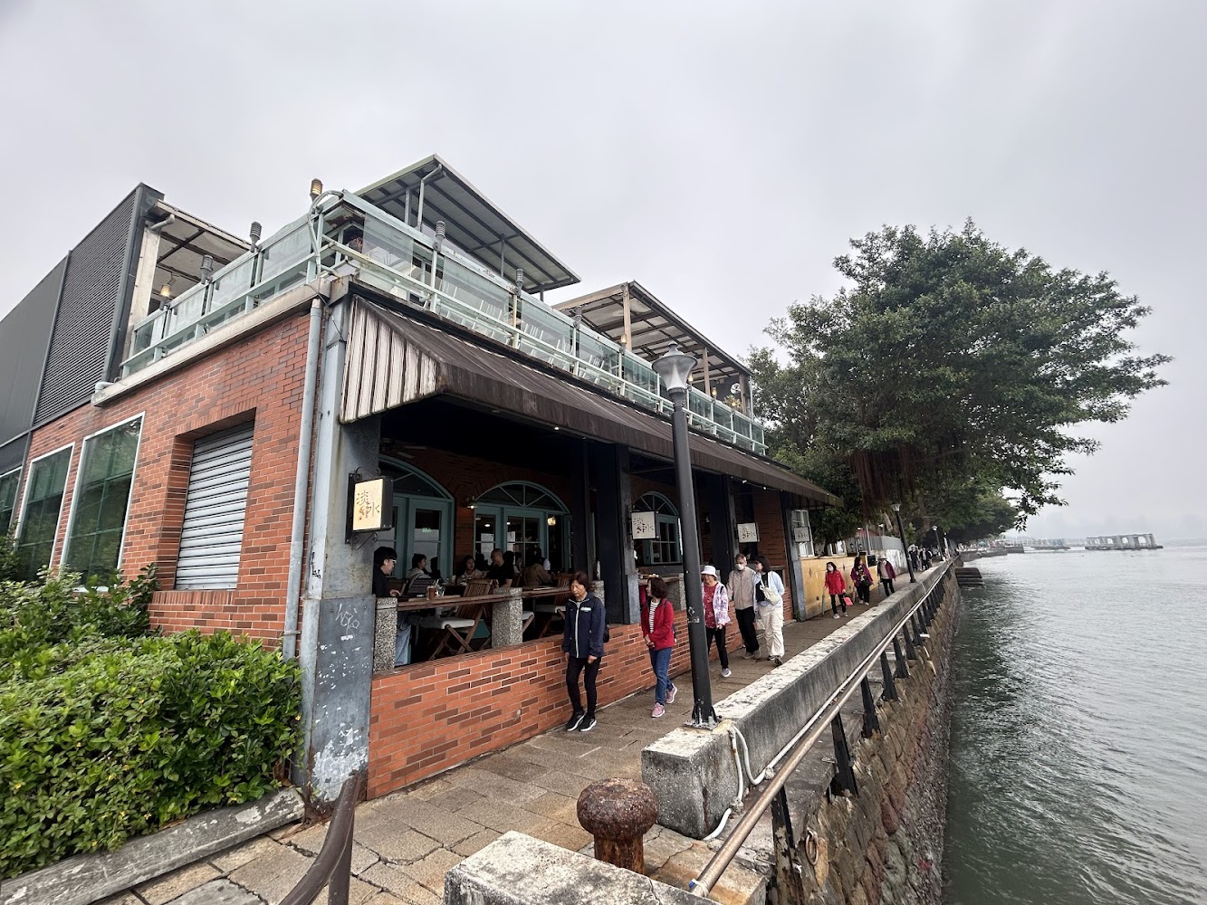 紅磚外牆餐廳位於河岸旁，裡面有顧客用餐，外面有人在步道散步