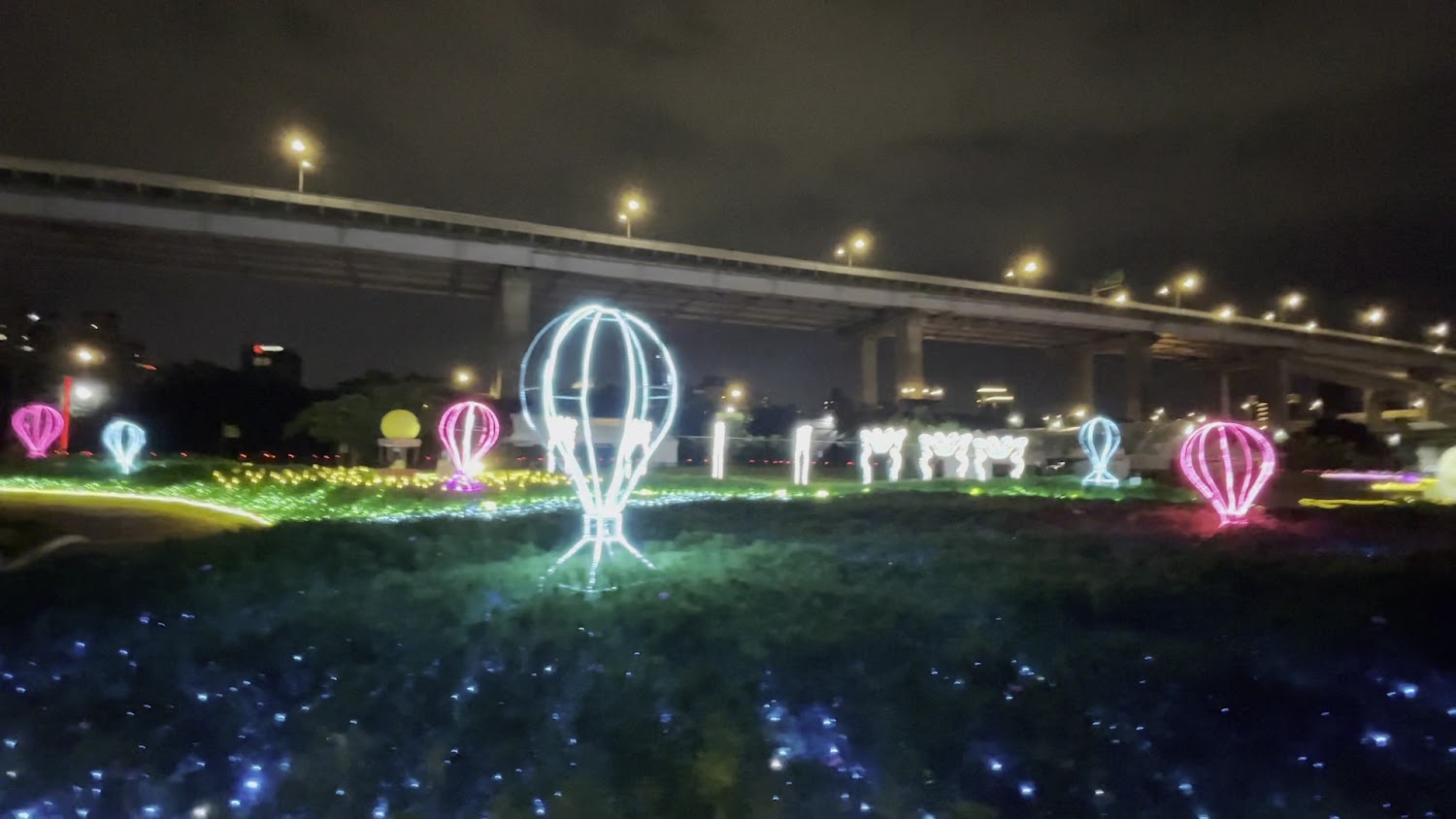 這張圖片主要展示了一個夜晚的燈飾花園，地上擺設許多熱氣球形狀的燈飾，並有五光十色的燈光點綴，背景有高架橋和夜空，營造浪漫氛圍。