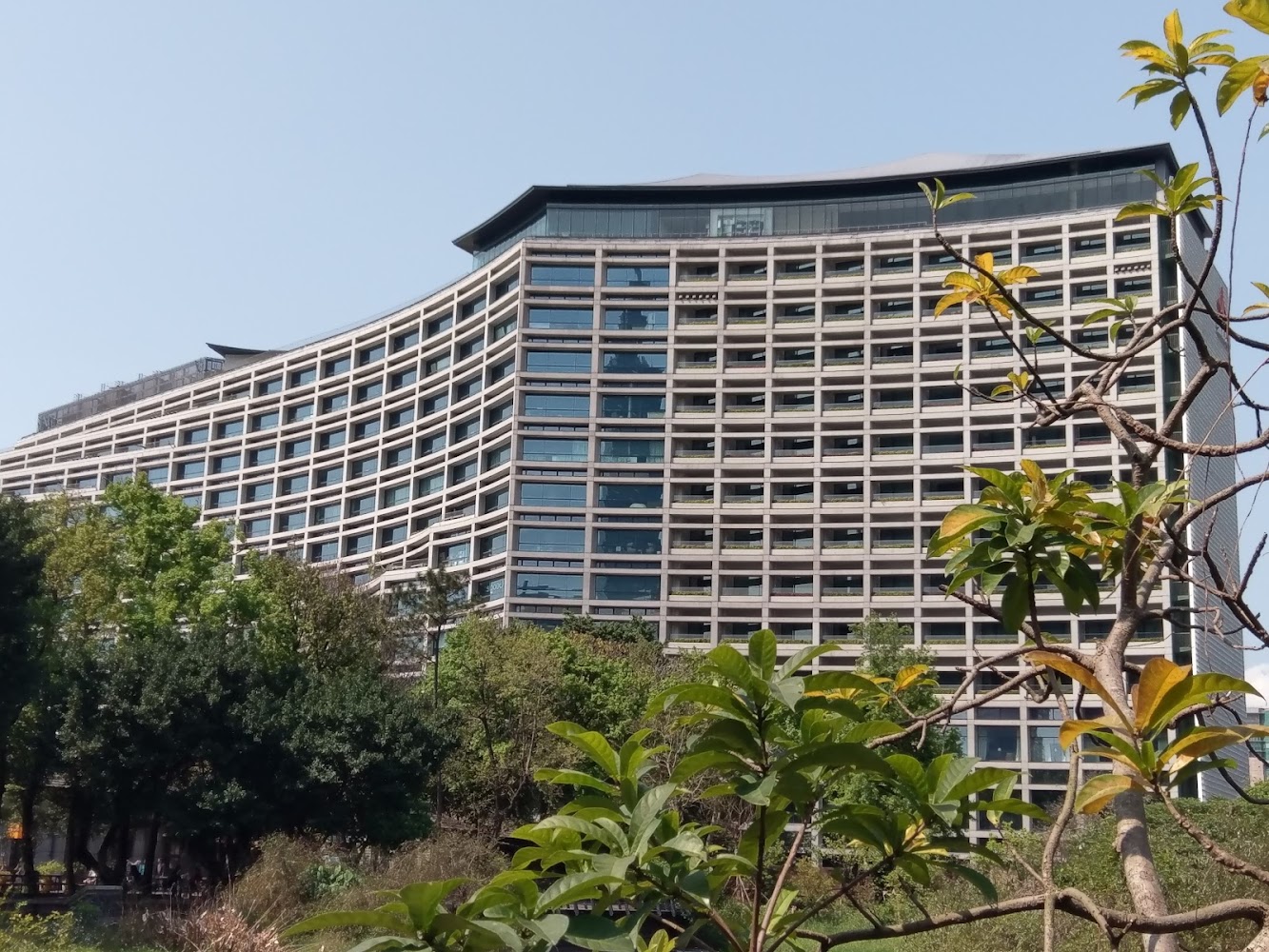 現代高樓建築，建築物外觀有曲線設計，周圍綠樹環繞，天氣晴朗，藍天與自然環境交織呈現都市與自然共融的景色。
