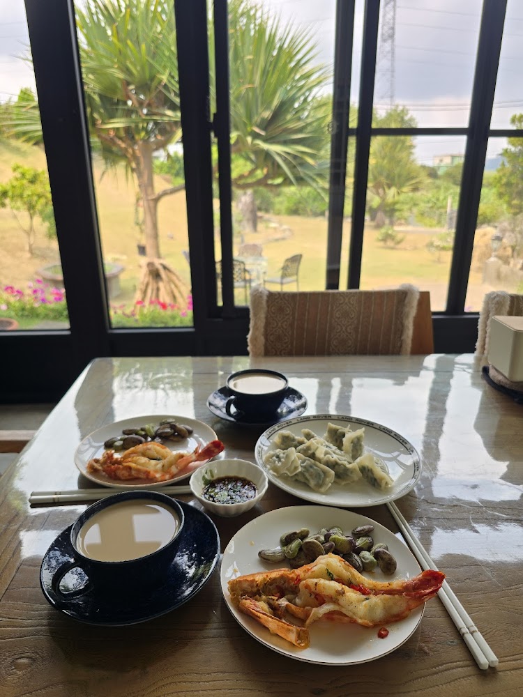這張圖片展示一張餐桌，上面有兩杯奶茶、兩盤蝦仁與豆類、一盤水餃和一小碗醬料。窗外是綠意盎然的花園景色，環境悠閒舒適。圖片沒有浮水印。
