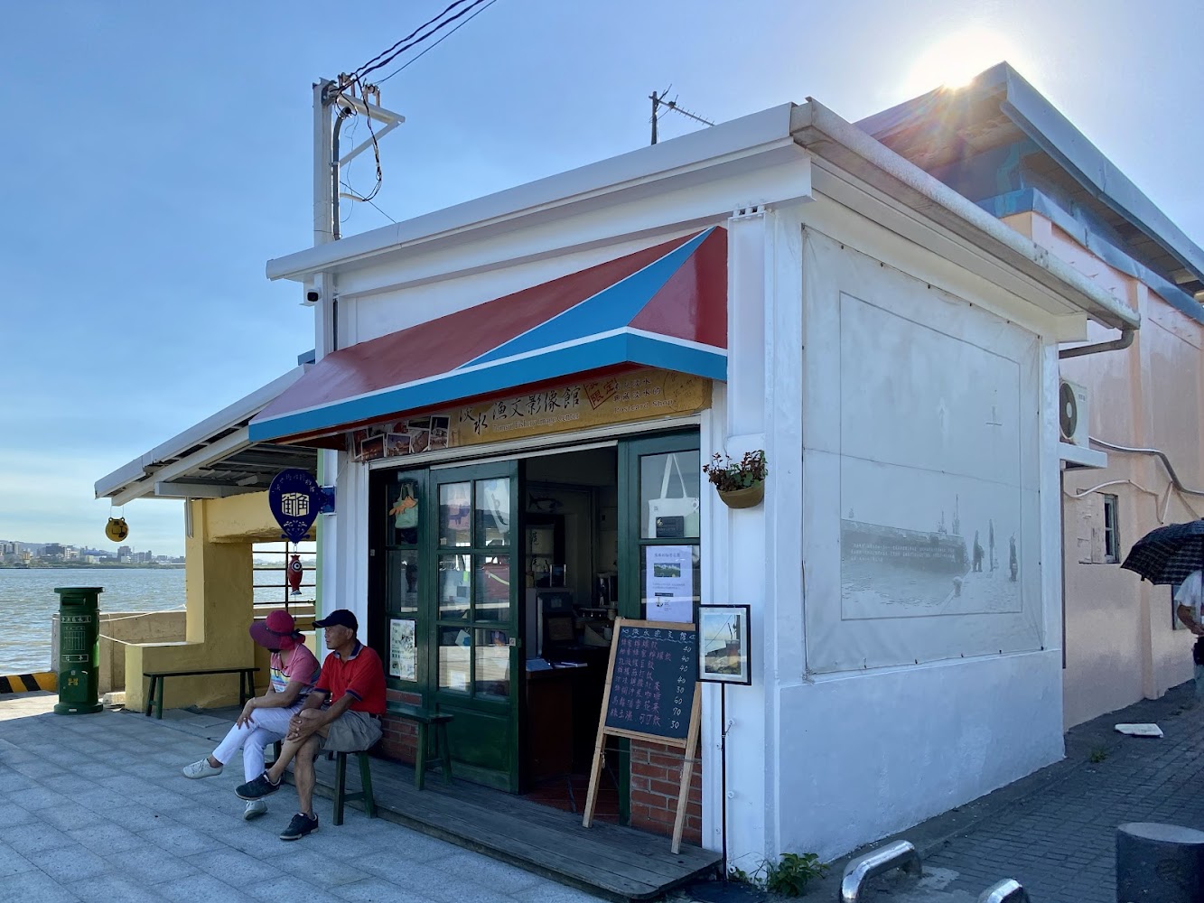 位於河邊的小店，有紅藍相間遮陽篷、玻璃門與門口休息的長者