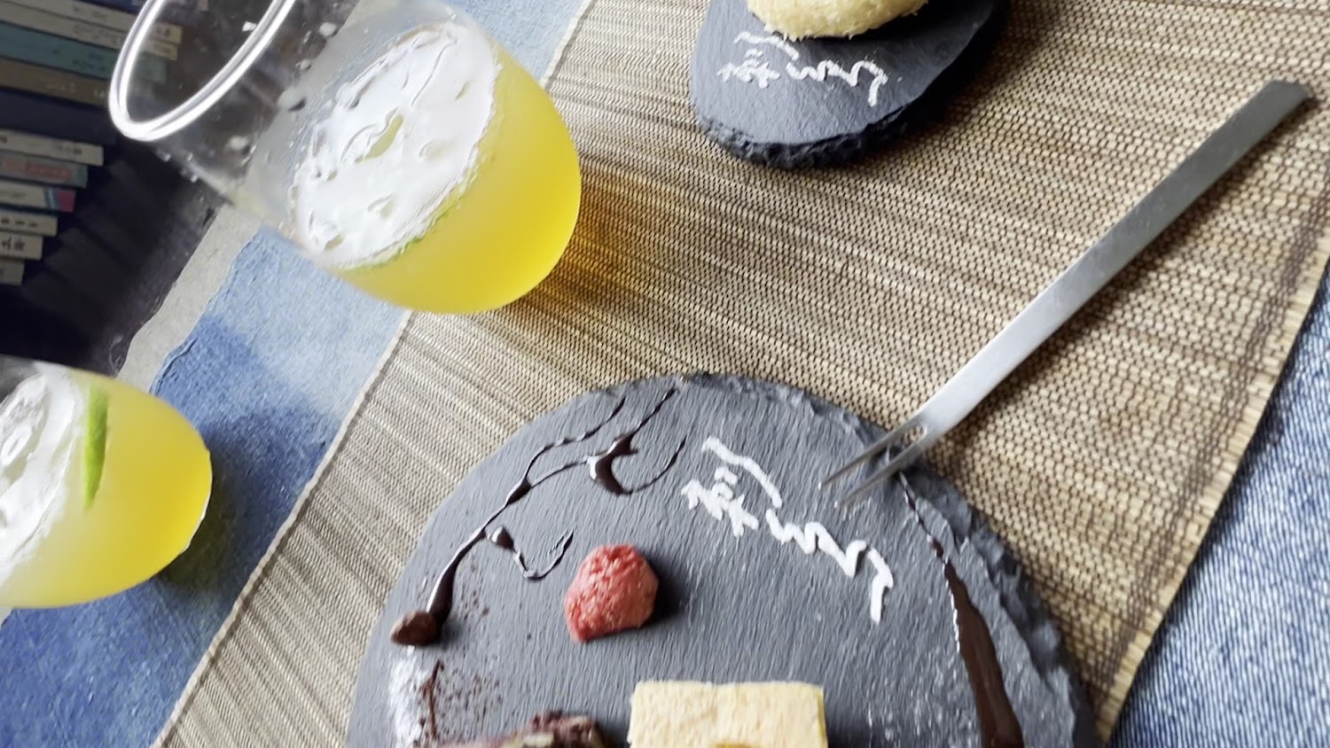這張圖片展示了兩杯加入檸檬片的黃色飲品，以及擺放有甜點和巧克力醬的黑色石板餐盤，整體搭配質感擺設於藤編桌墊上。