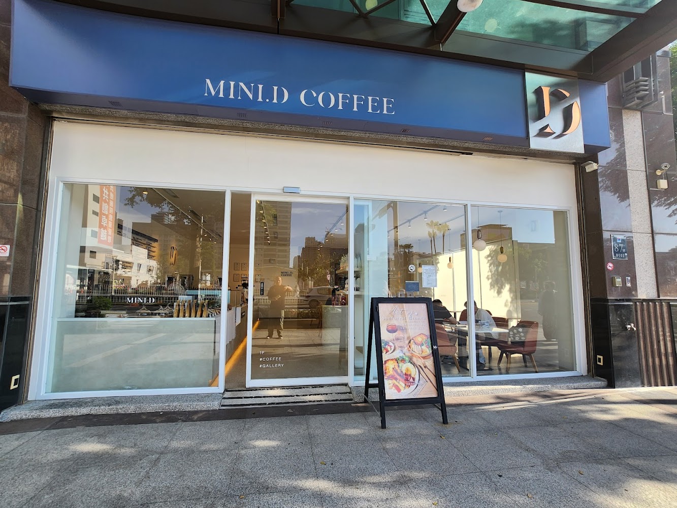 這是一家名為MINI.D COFFEE的咖啡廳外觀,玻璃門前放有一個展示美食的廣告牌,透過玻璃可以看到店內座位和顧客,環境現代明亮。