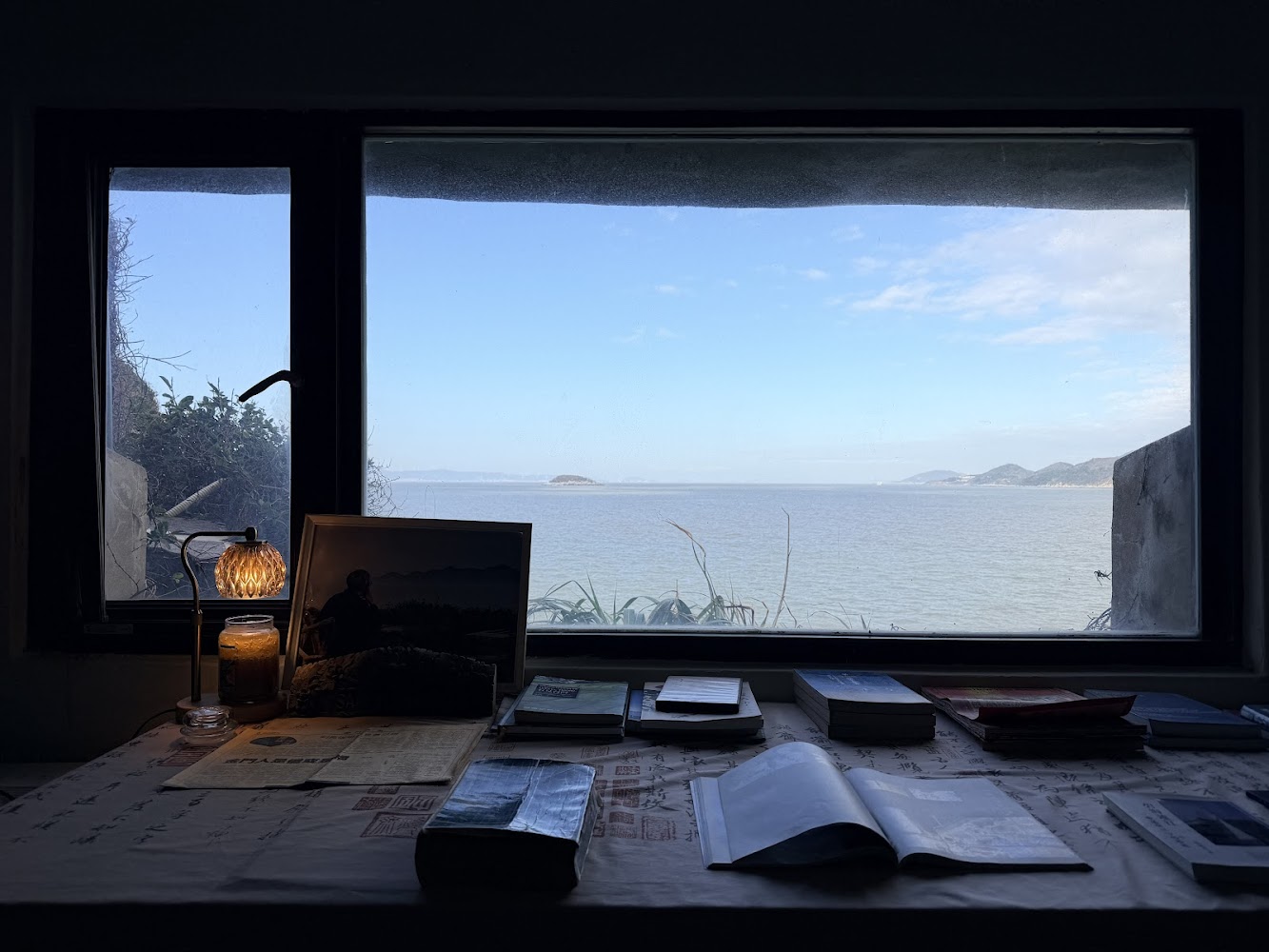 這張圖片展示一張朝向窗外大海的書桌，桌上擺放著書本、雜誌、相框與檯燈。窗外可見藍天與海景，遠方有山脈和小島，環境寧靜宜人。