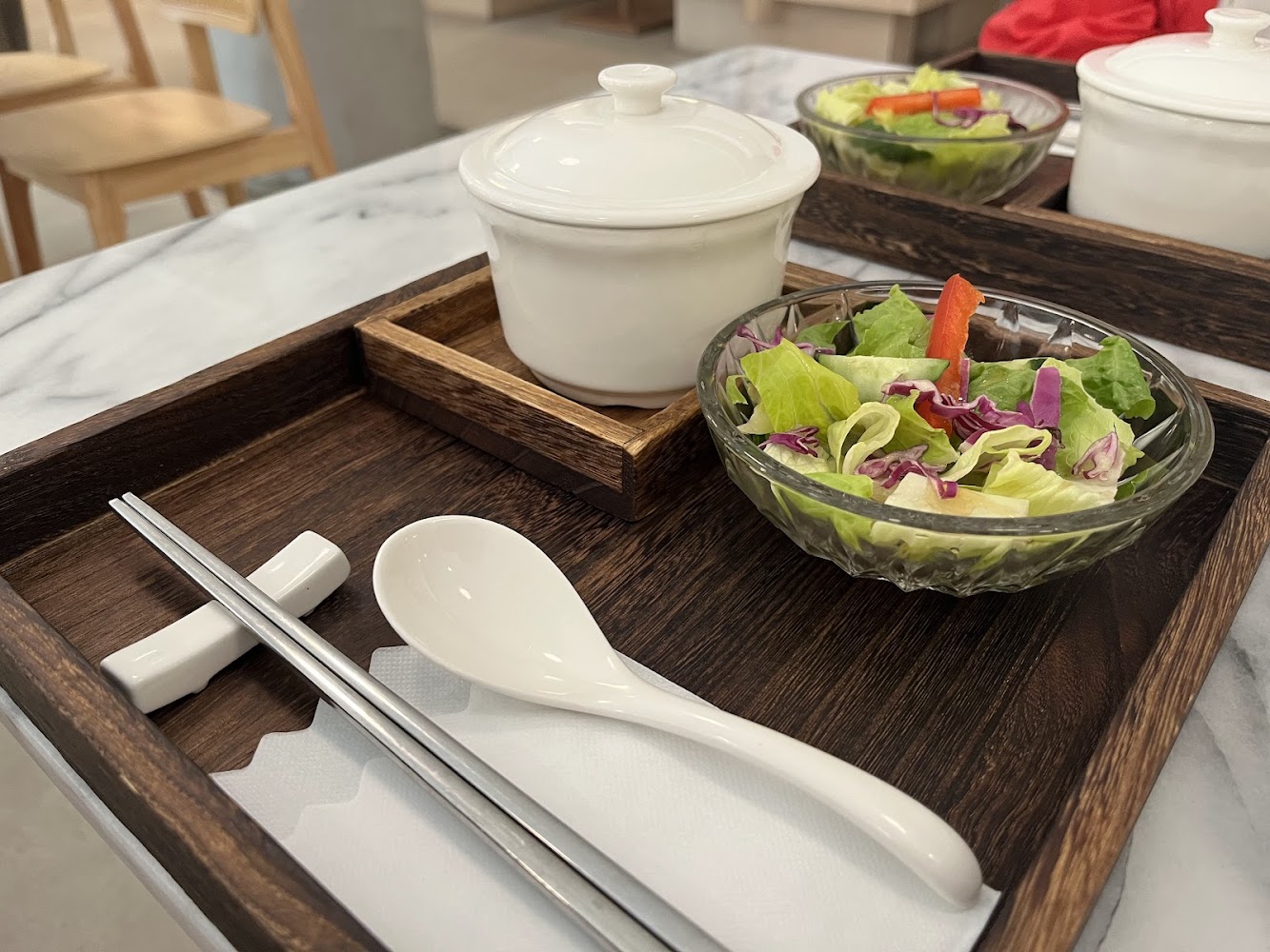 這張圖片展示了一套餐具，包含一盤新鮮蔬菜沙拉、一個陶瓷湯碗、筷子與湯匙，整齊地擺放在木質托盤上，給人簡潔清新的用餐感覺。