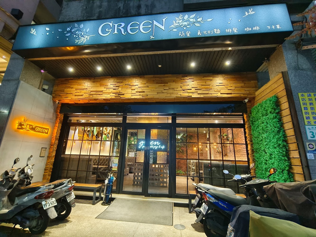 這是一家名為GREEN的餐廳門口，外觀有植栽裝飾及木質設計，門口停有數輛機車，氣氛溫馨。招牌標示提供簡餐、義大利麵、排餐、咖啡及下午茶。