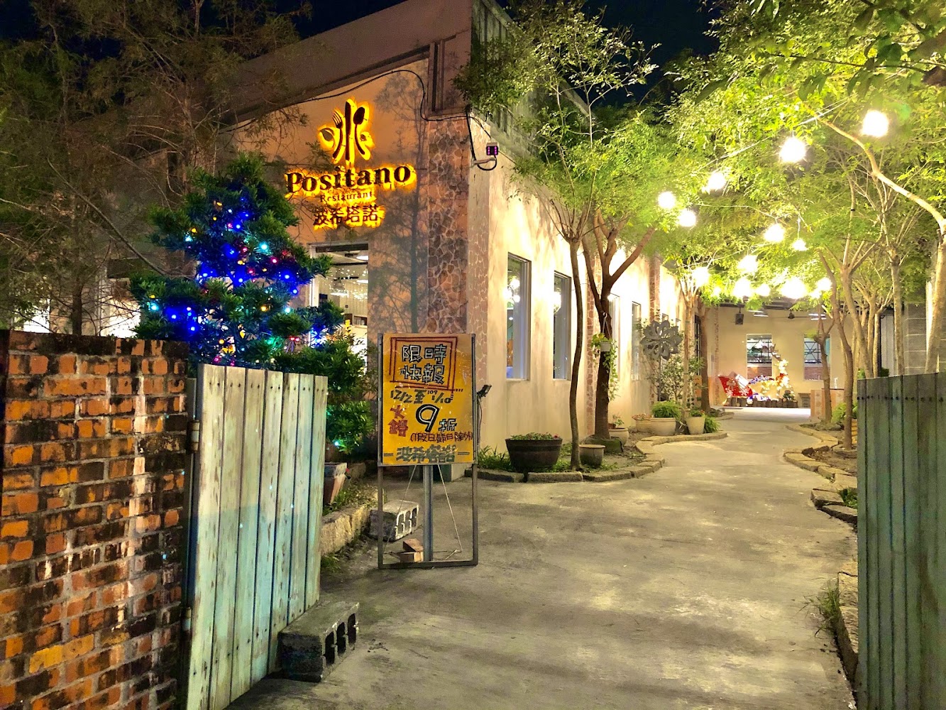 這張圖片主要展現一家名為Positano的餐廳夜景，入口兩側有綠樹與彩燈裝飾，氛圍溫馨，有告示牌和燈光點綴，通道深處還能看到聖誕裝飾品。