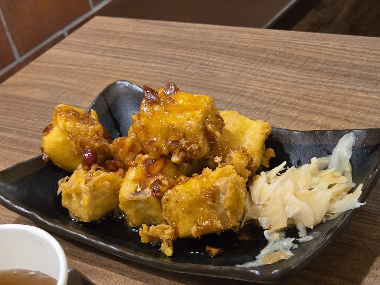 這張圖片主要有一盤酥炸豆腐，淋上醬汁，旁邊搭配一些醃製薑片，擺放在黑色陶瓷盤上，桌面為木質紋路。