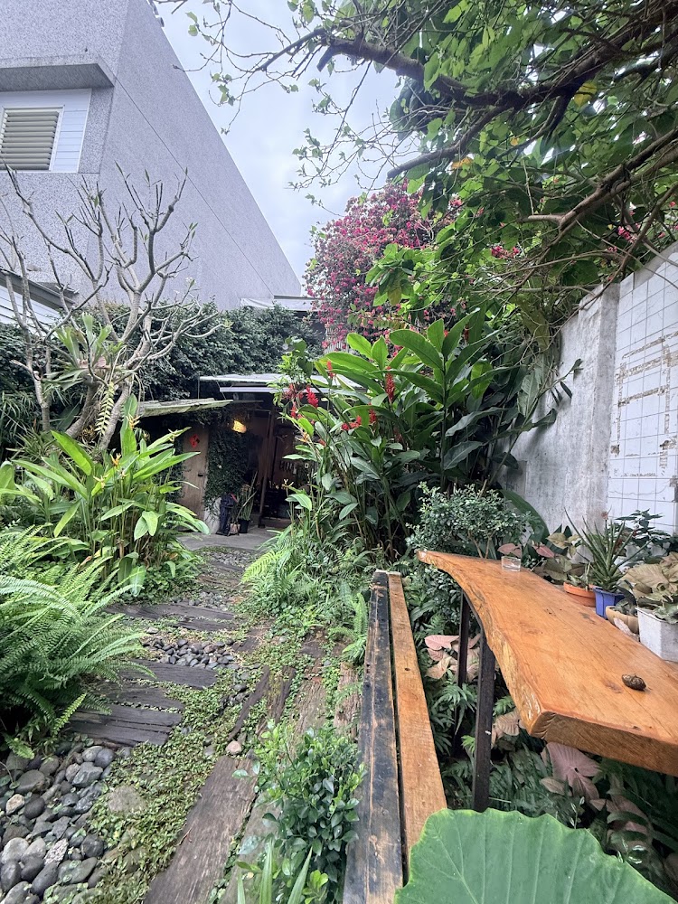 這張圖片展示了一個綠意盎然的戶外小庭院，中央有石板與木棧道通往一間小屋。右側有木頭長桌和長椅，周圍環繞各種熱帶植物及花草，景色自然舒適。